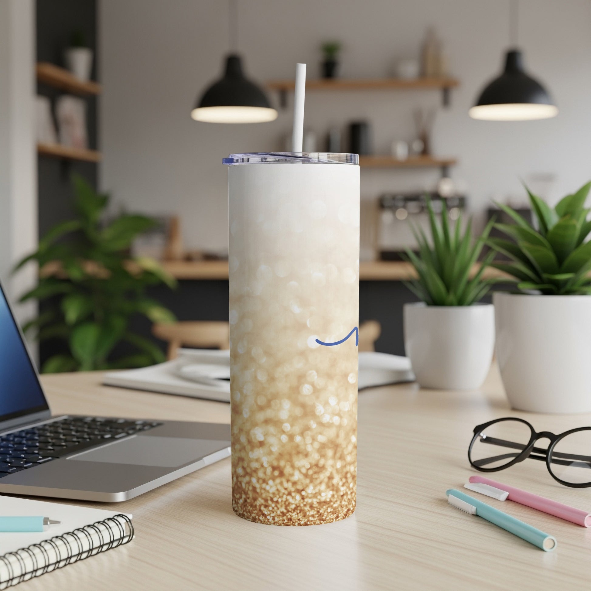 Glittery gold tumbler with blue swirl logo on desk with laptop and pens.