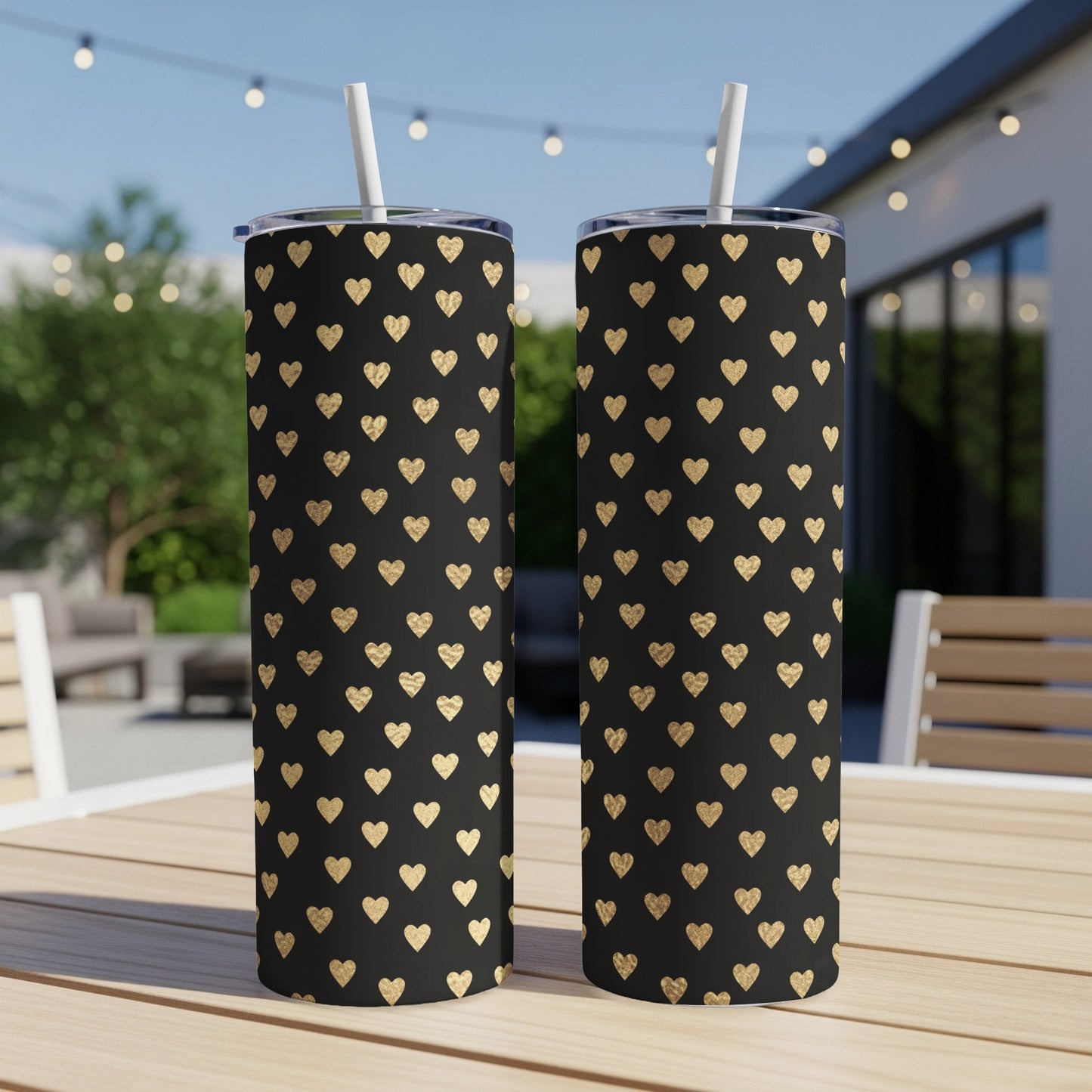 Two black tumblers with a gold heart pattern and straws sit on a wooden table.
