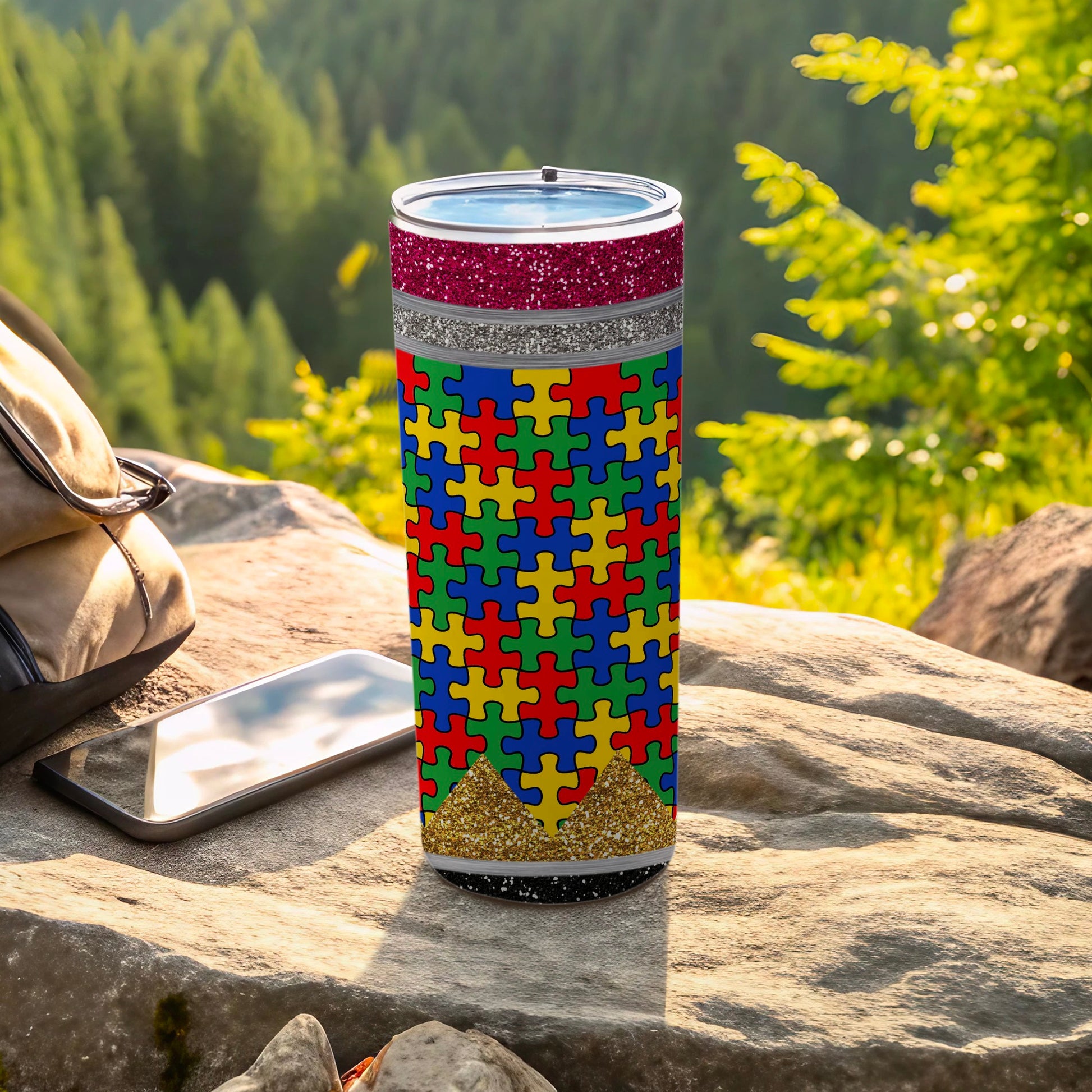 Autism awareness puzzle tumbler with glitter accents on outdoor rock surface.