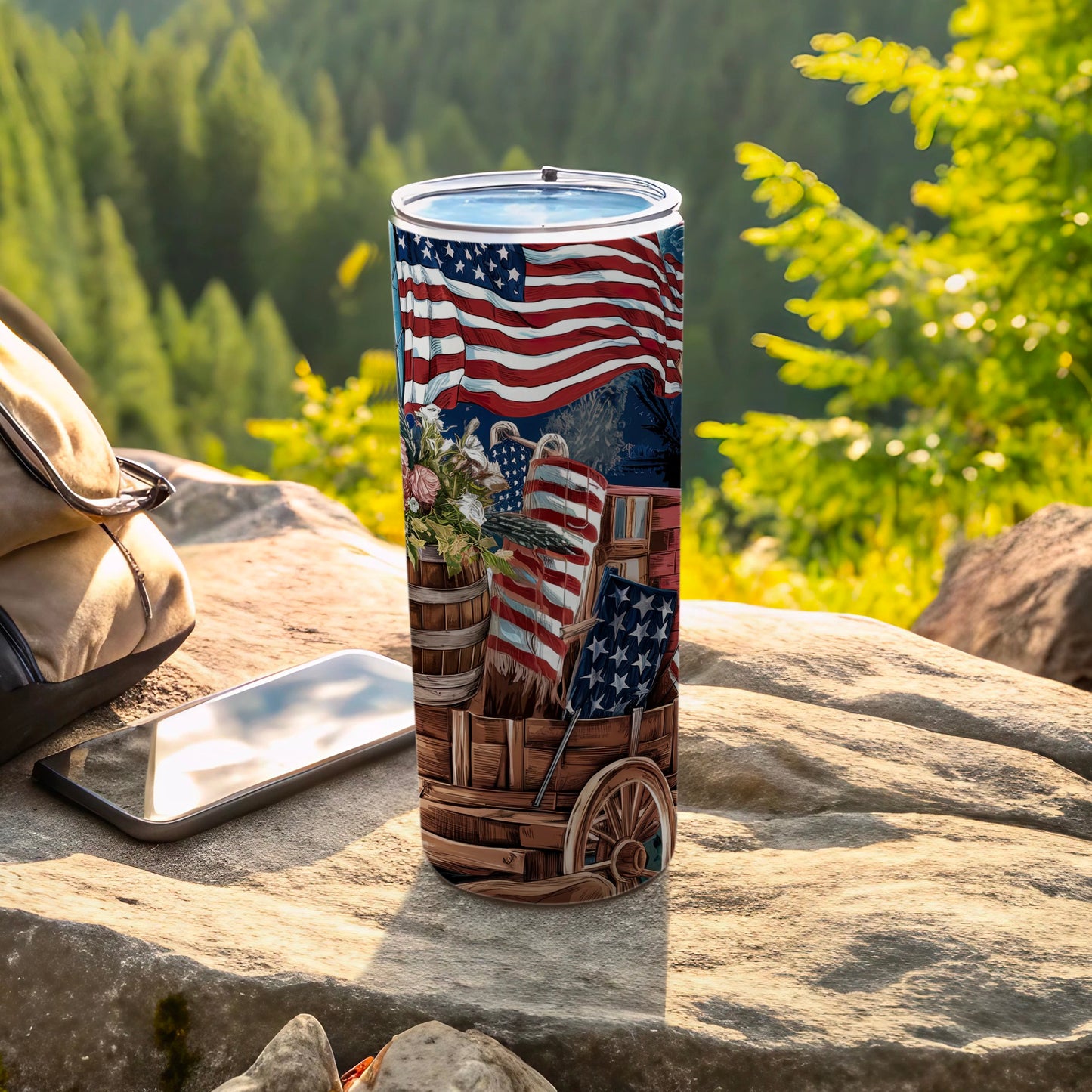 Tall tumbler with American flag design and wagon, on rock with phone and backpack.