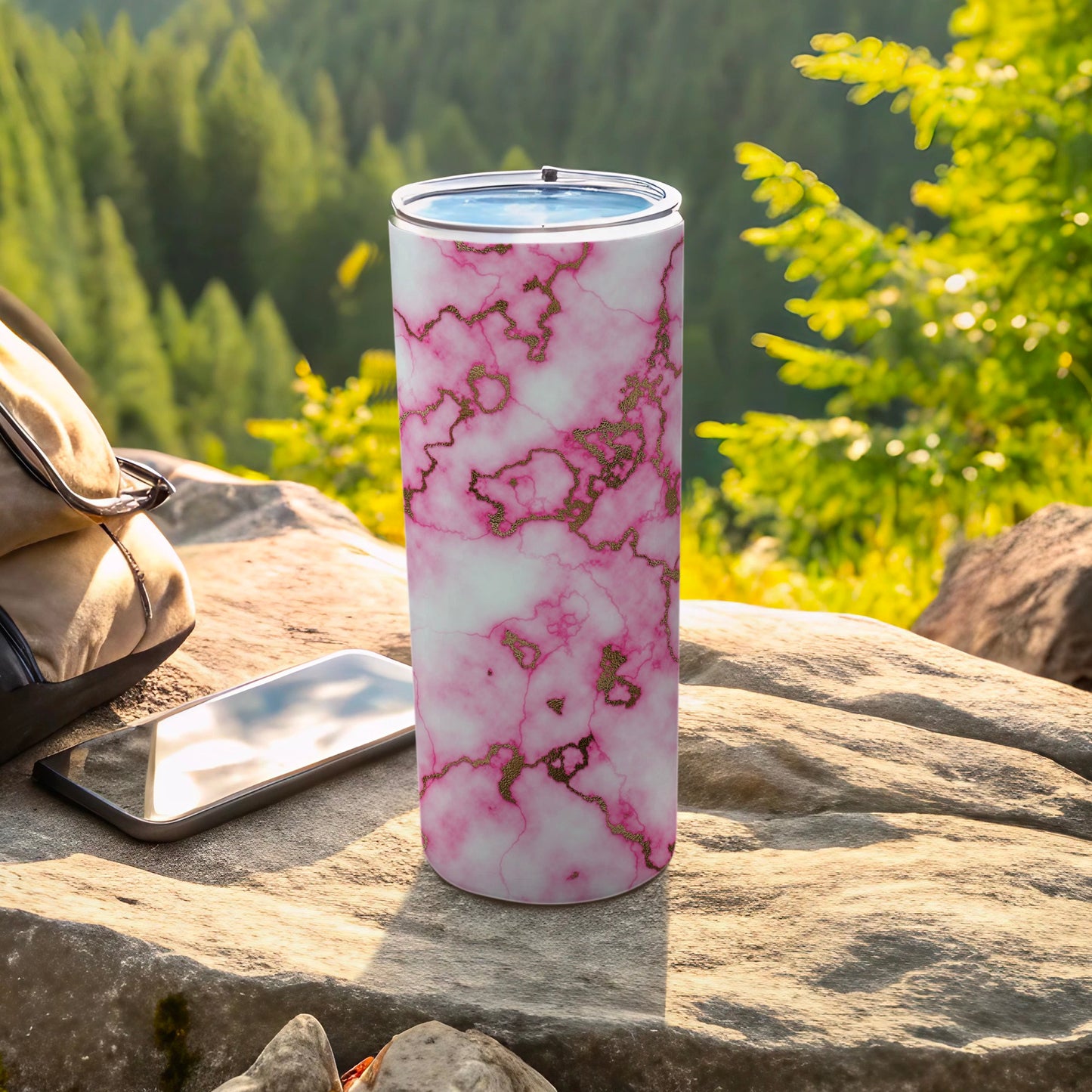 Pink marble tumbler with gold accents on a rock next to a phone and backpack.