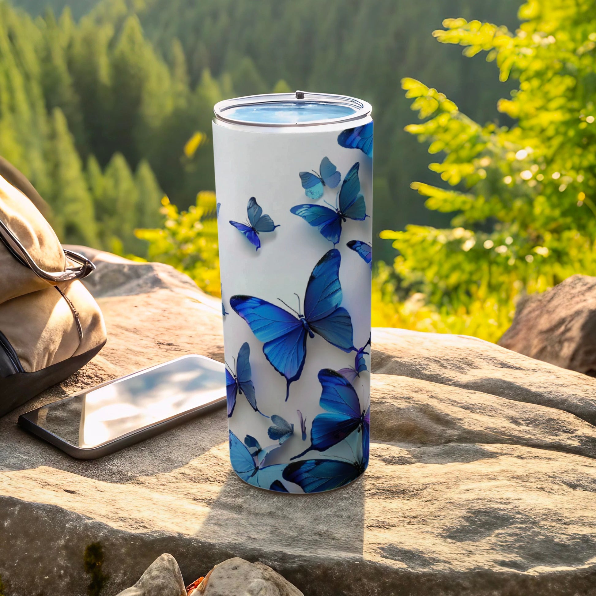 White tumbler with blue butterflies, resting on rocks with a backpack and phone.