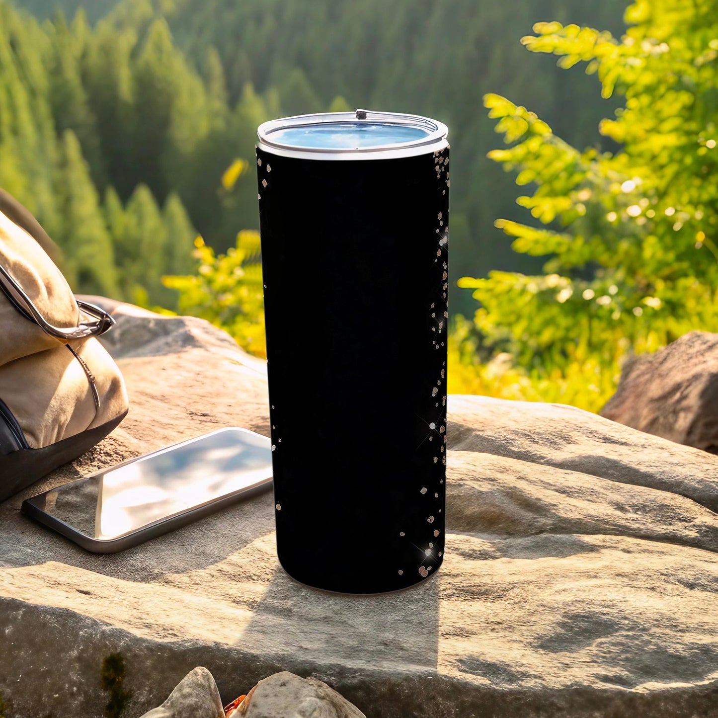 Black tumbler with glitter speckles, resting on rock with phone and bag.