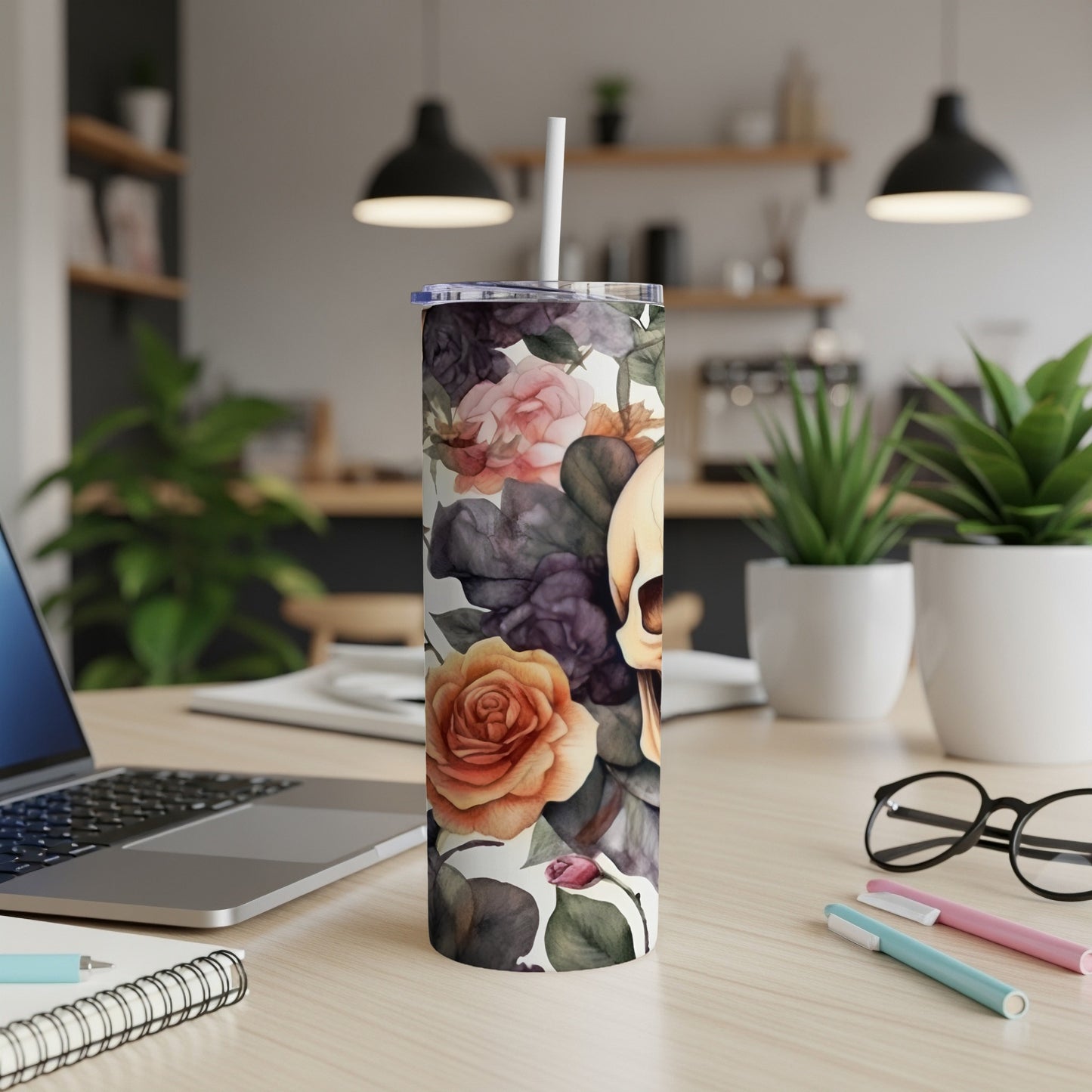 Floral skull tumbler with a straw on a desk with a laptop and pens.