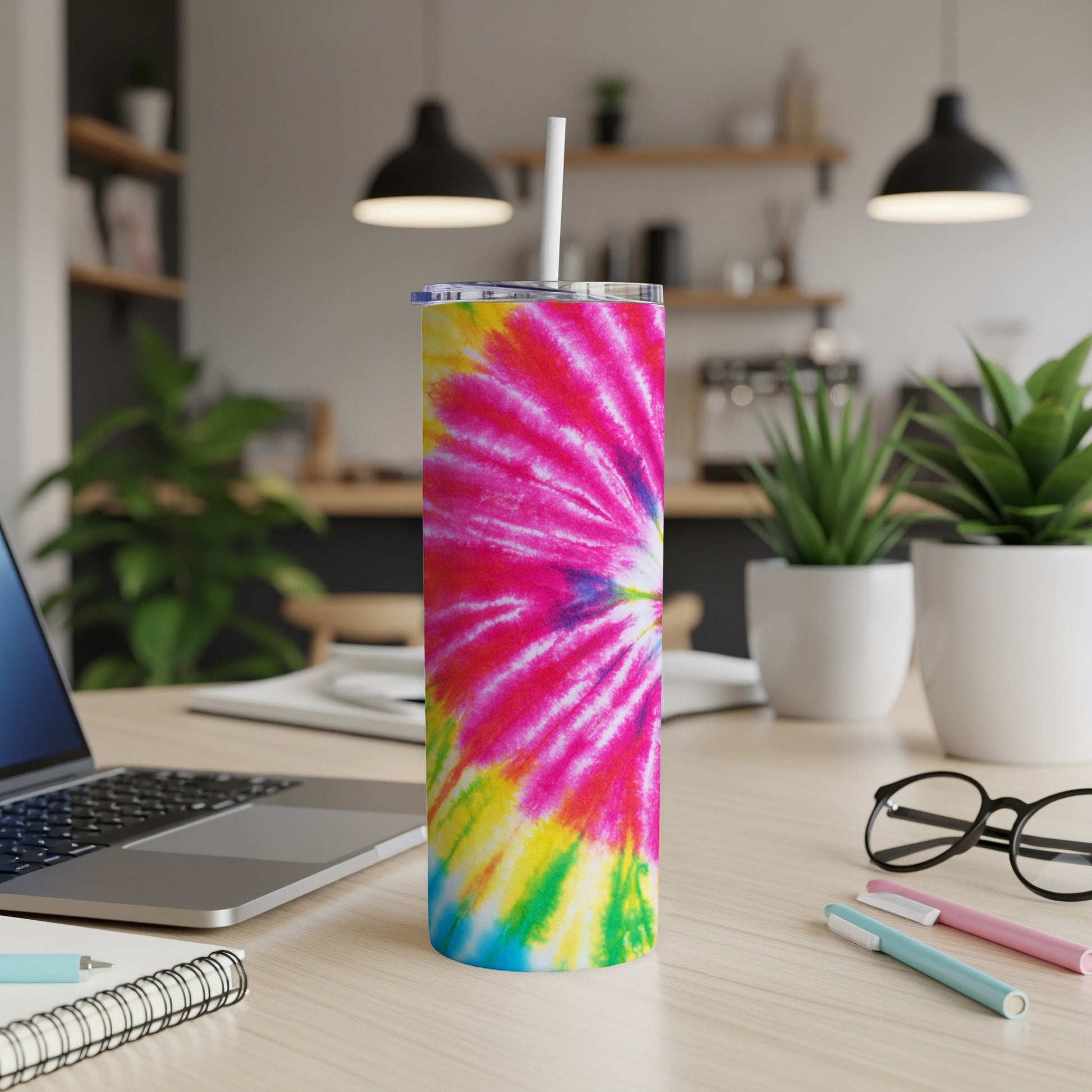 Tie-dye tumbler with straw on desk with laptop and pens.