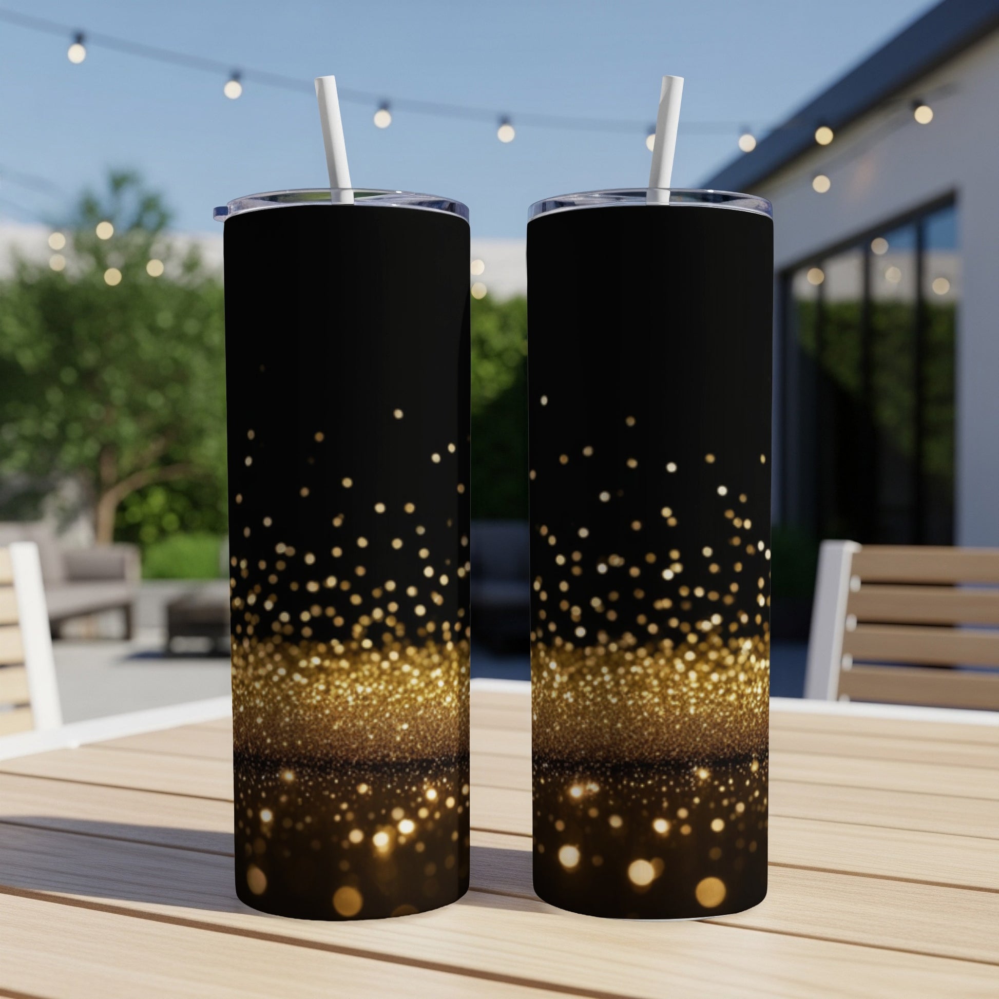 Two black tumblers with gold glitter gradient pattern, with straws, on outdoor table.