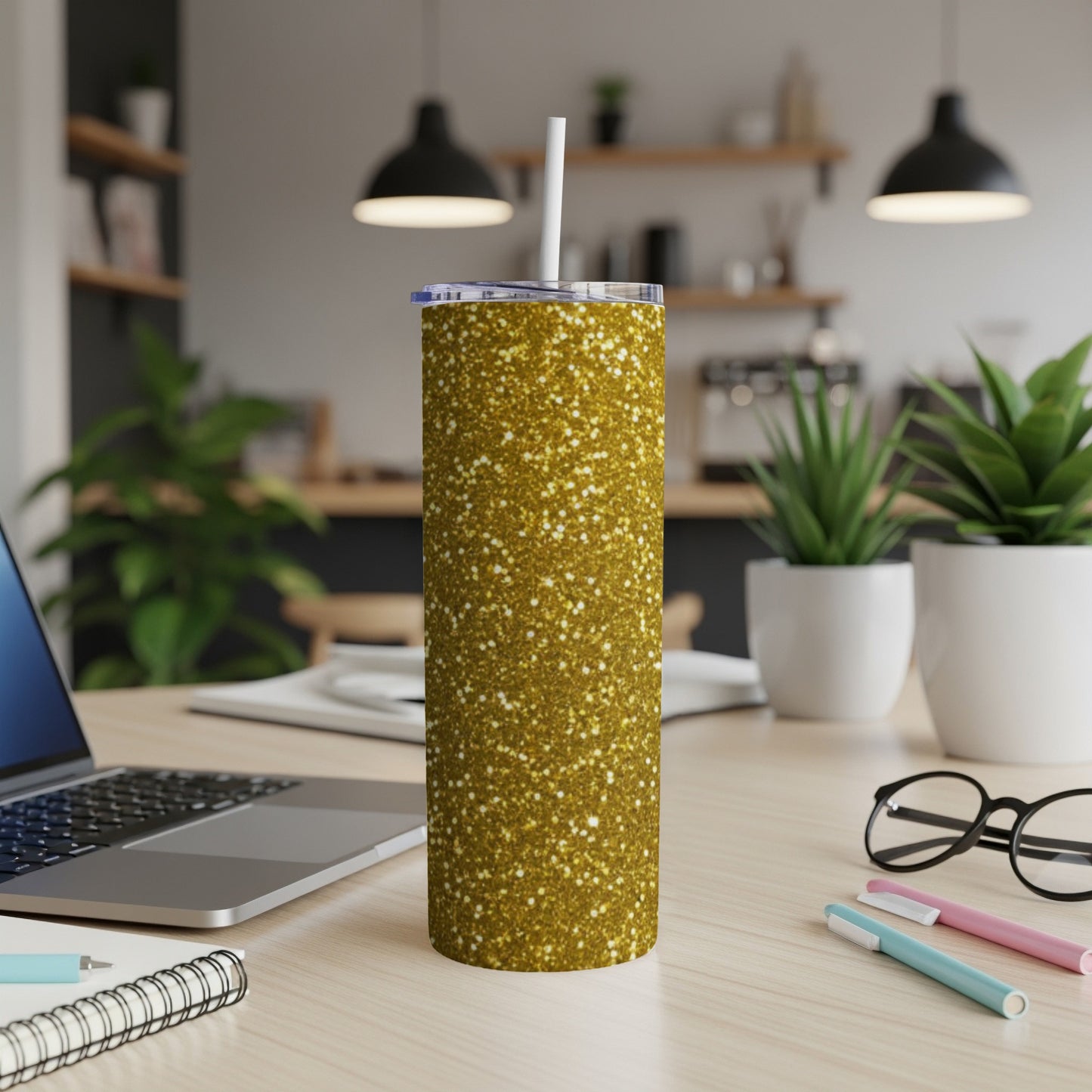 Gold glitter tumbler with straw on a wooden desk with laptop and plants.