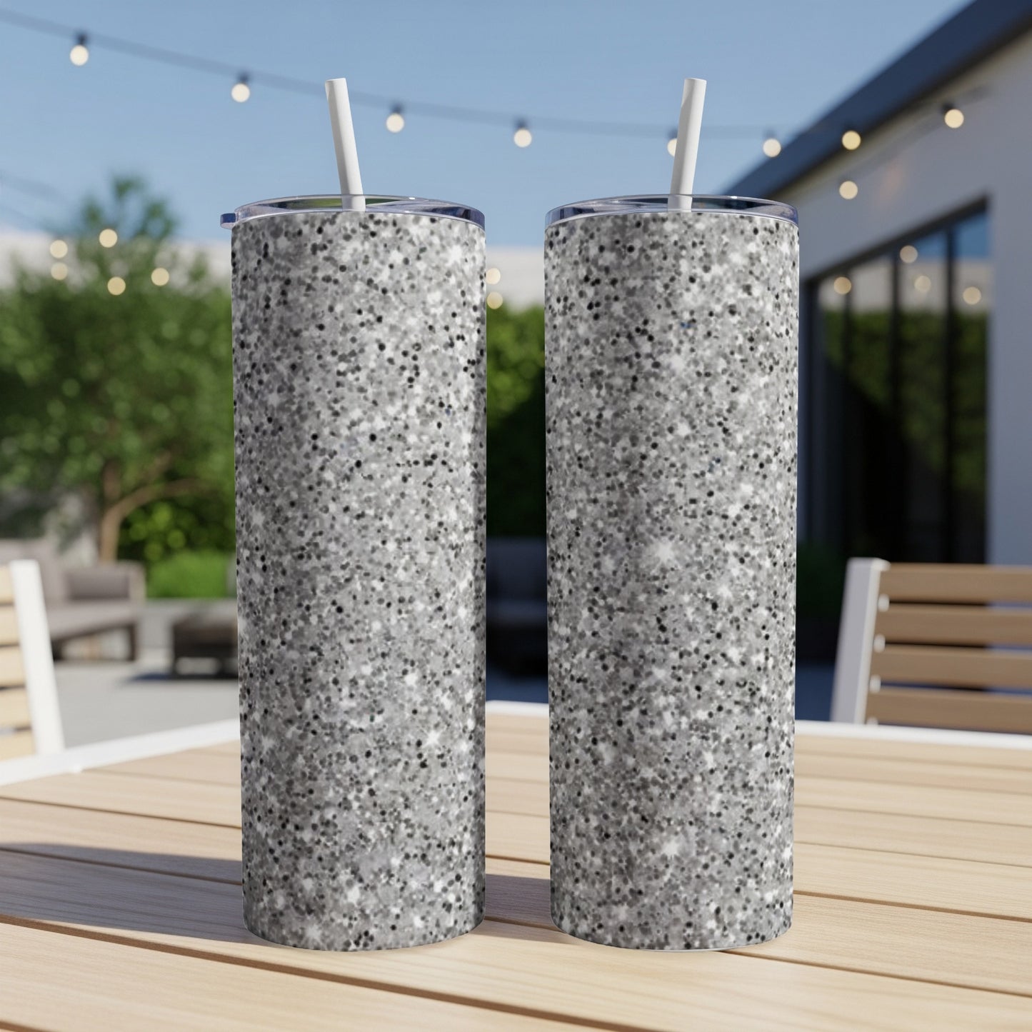 Two silver glitter tumblers with straws on a wooden table outdoors.