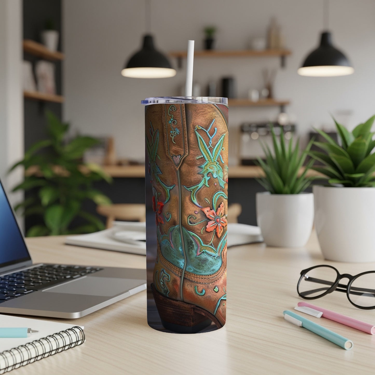 Brown leather cowboy boot tumbler with floral design and straw on a desk.