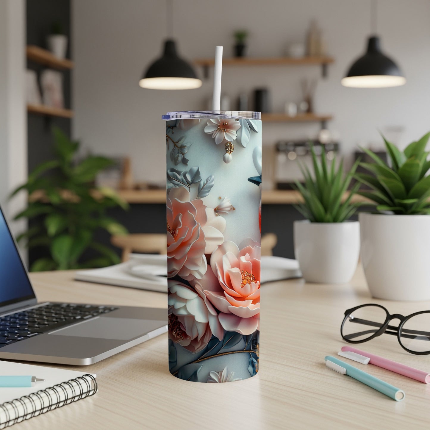 Floral tumbler with straw on a wooden desk with laptop and pens.