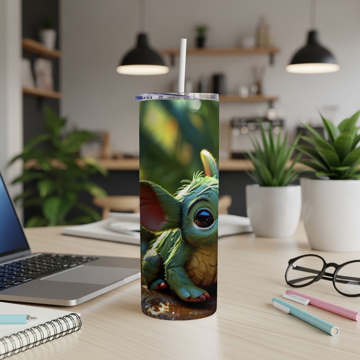 Green creature with big eyes on a tumbler, with laptop and pens.