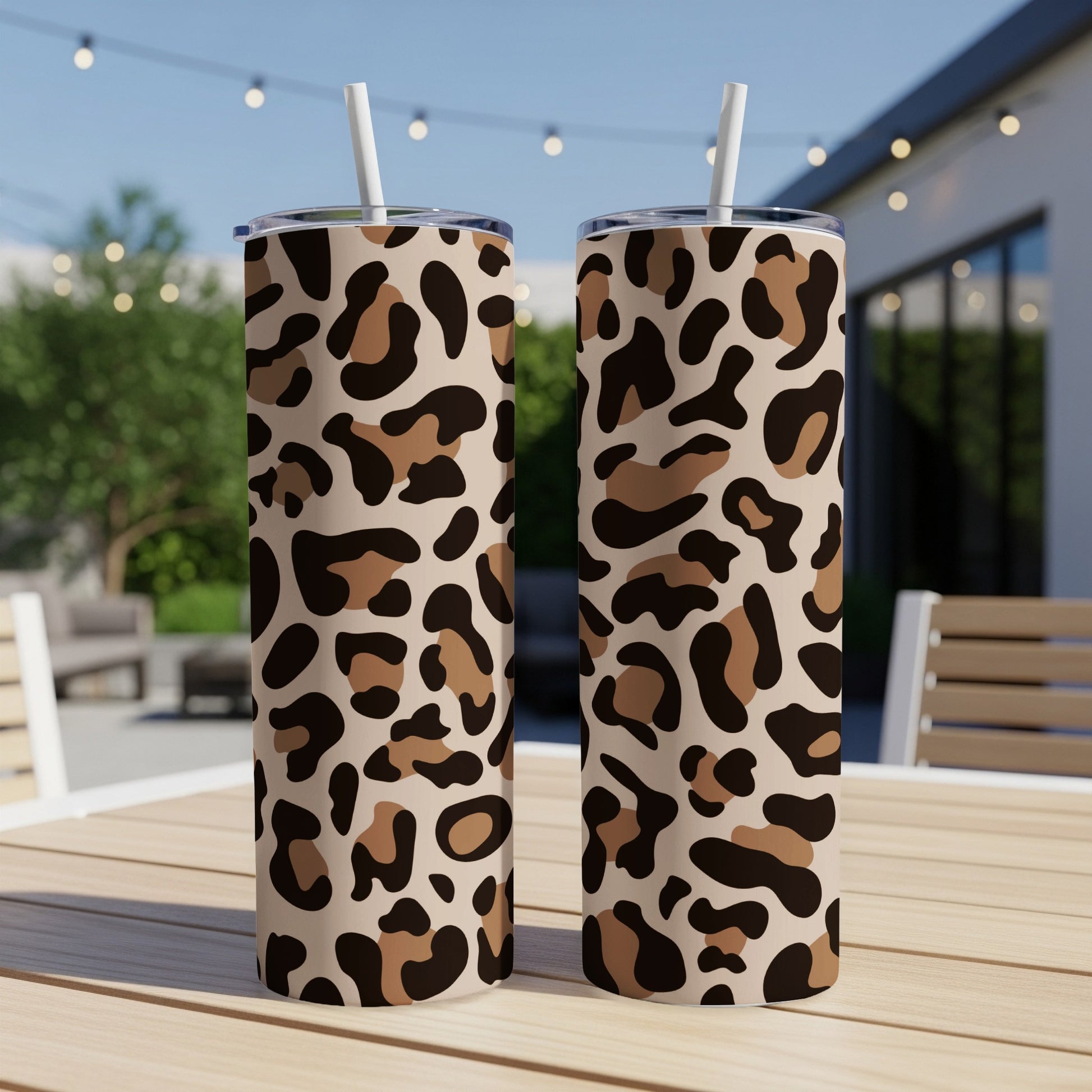Two leopard print tumblers with straws on a wooden table outdoors.