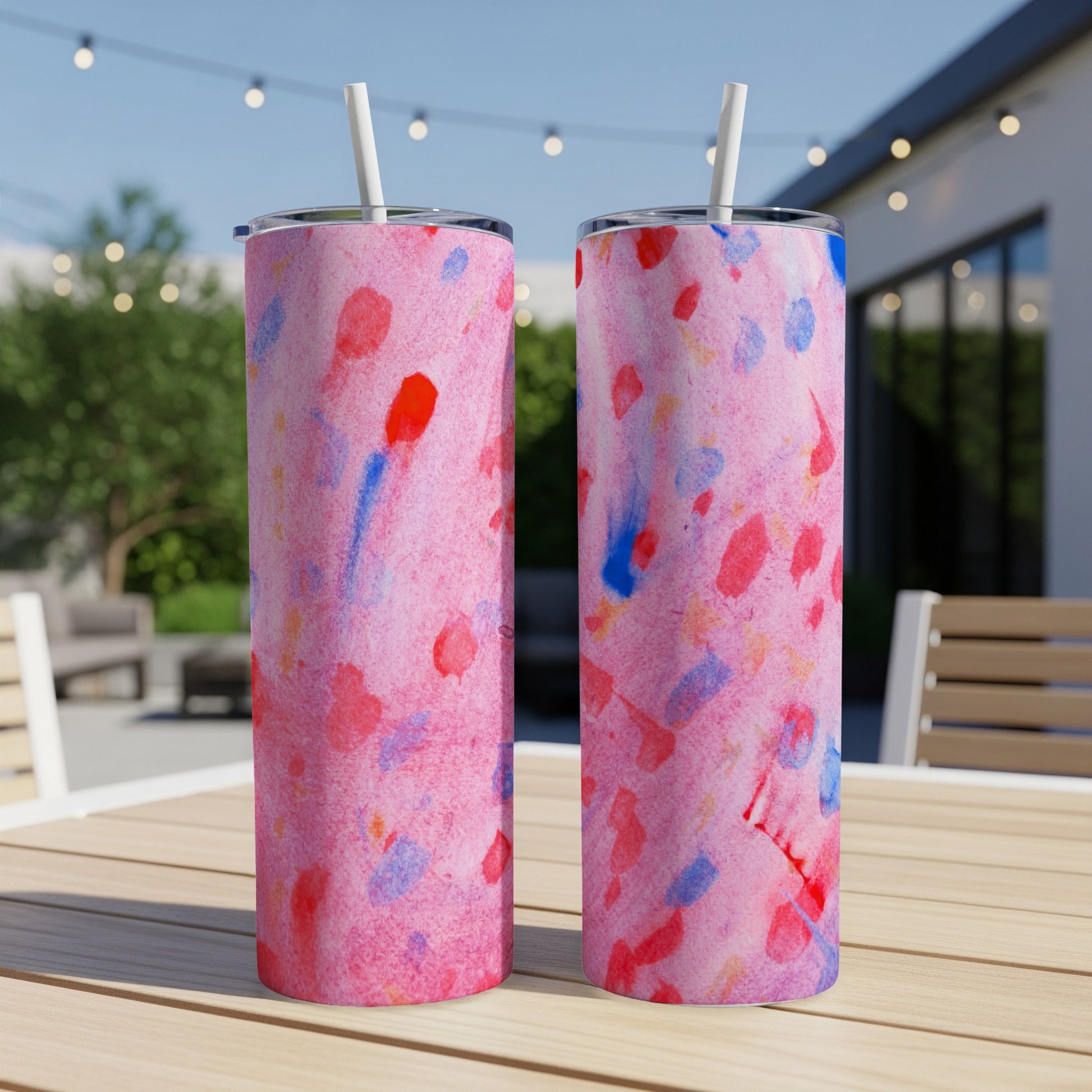 Two pink tumblers with red and blue abstract paint splatters, on a table outdoors.