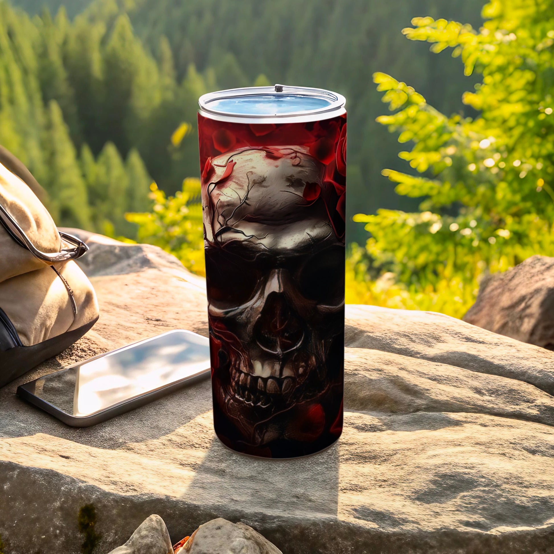 Tumbler with skull and rose design on a rock with phone and bag.