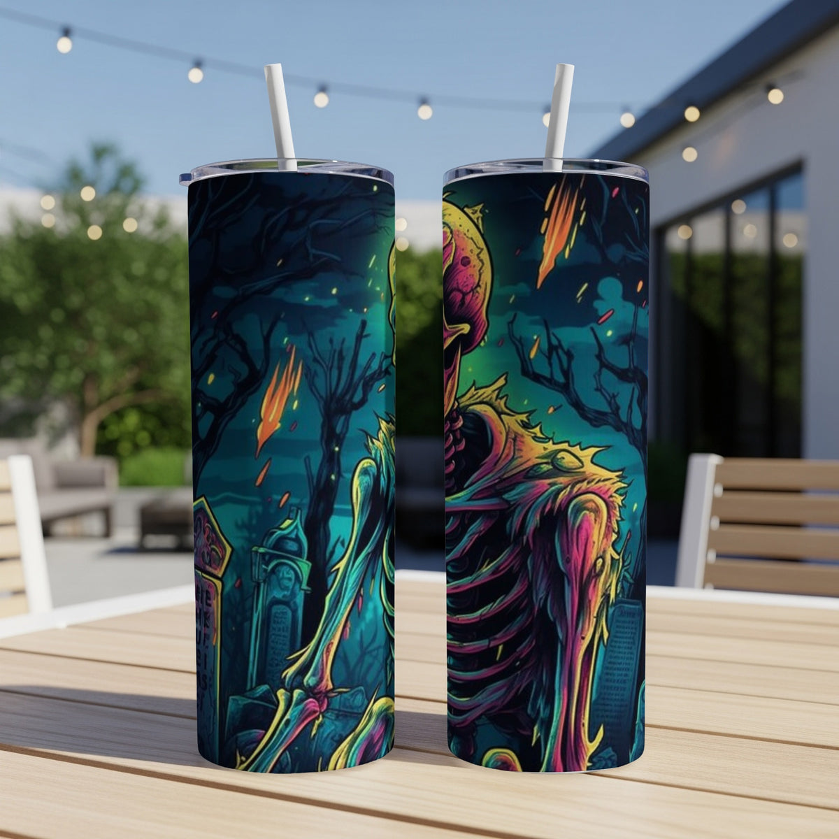 Two tumblers with a colorful skeleton and graveyard art, with straws, on a table outdoors.