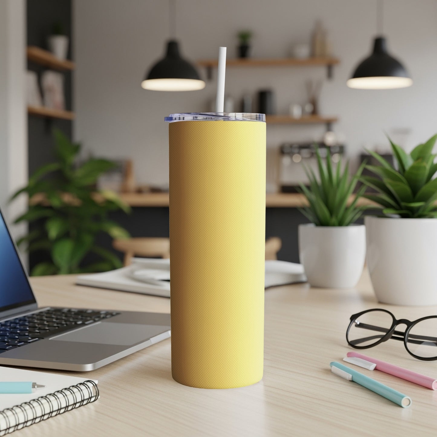 Yellow tumbler with straw on a desk next to a laptop and pens.