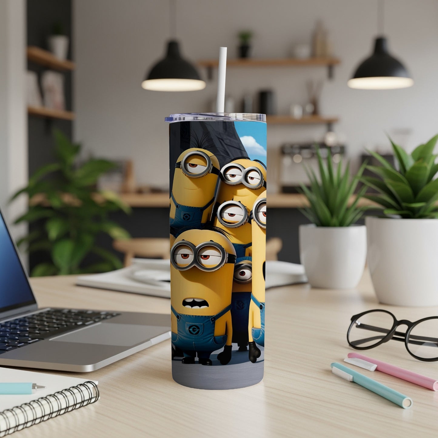 Minions characters on a tumbler with a straw, on a desk with a laptop and pens.