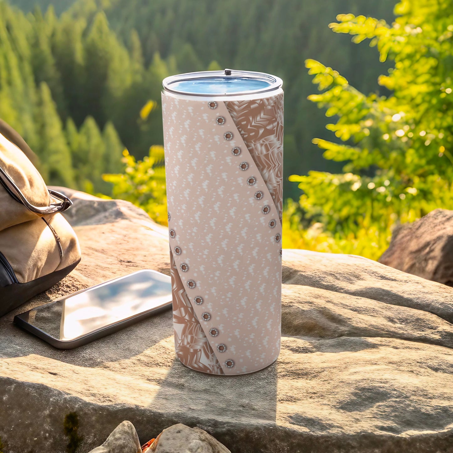 Tan tumbler with brown floral pattern on a rock, with phone and backpack nearby.