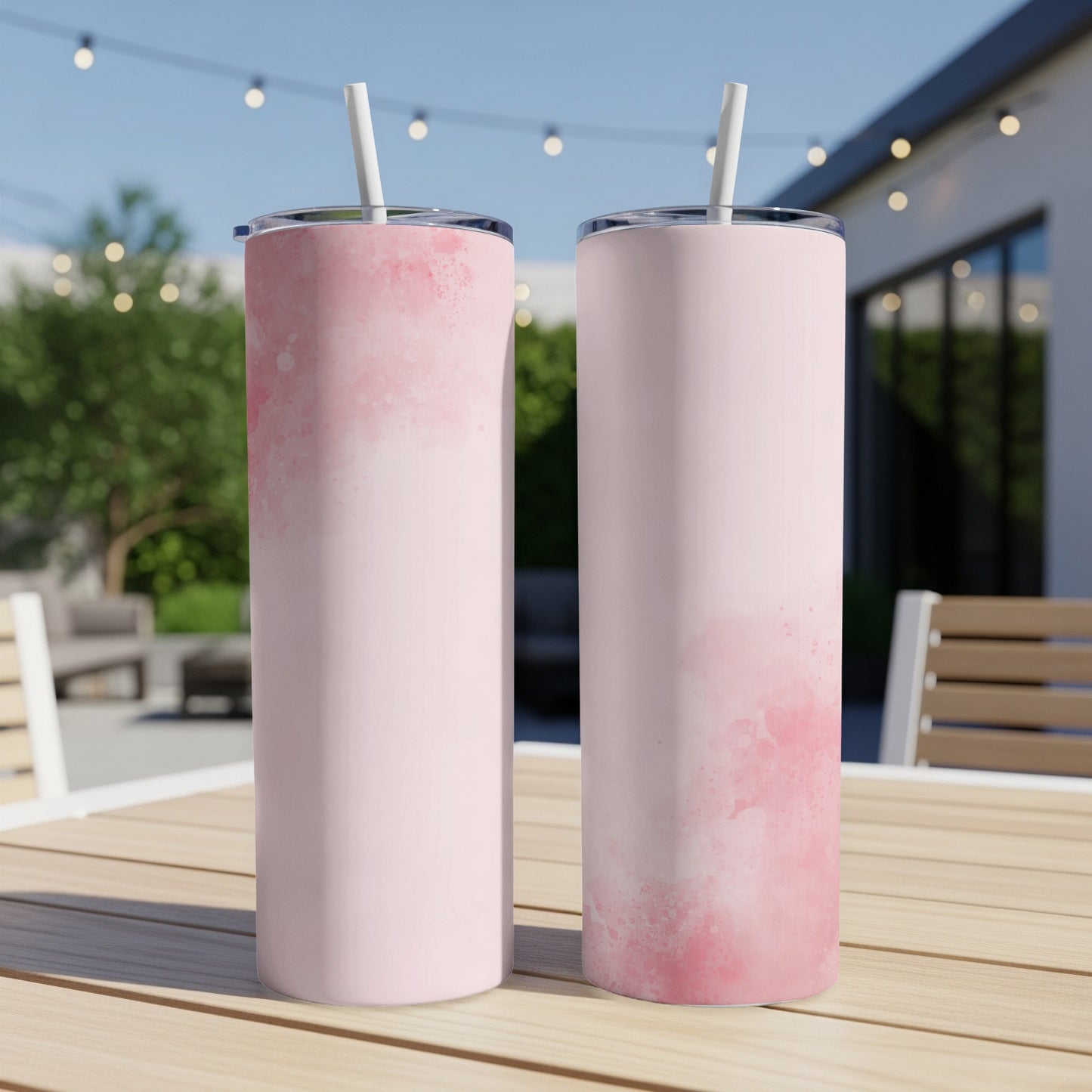 Two pink watercolor-splattered skinny tumblers with straws on a wooden table outdoors.