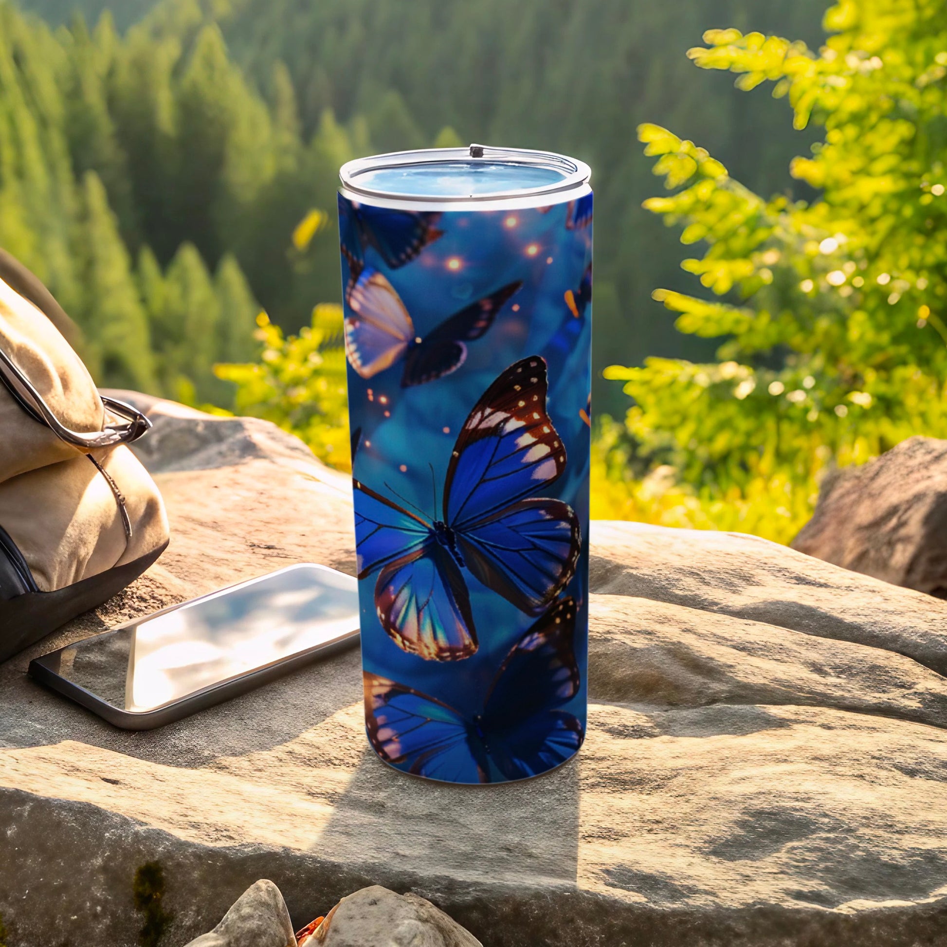 Blue butterflies on a cylindrical tumbler placed on rocks with a backpack and phone.