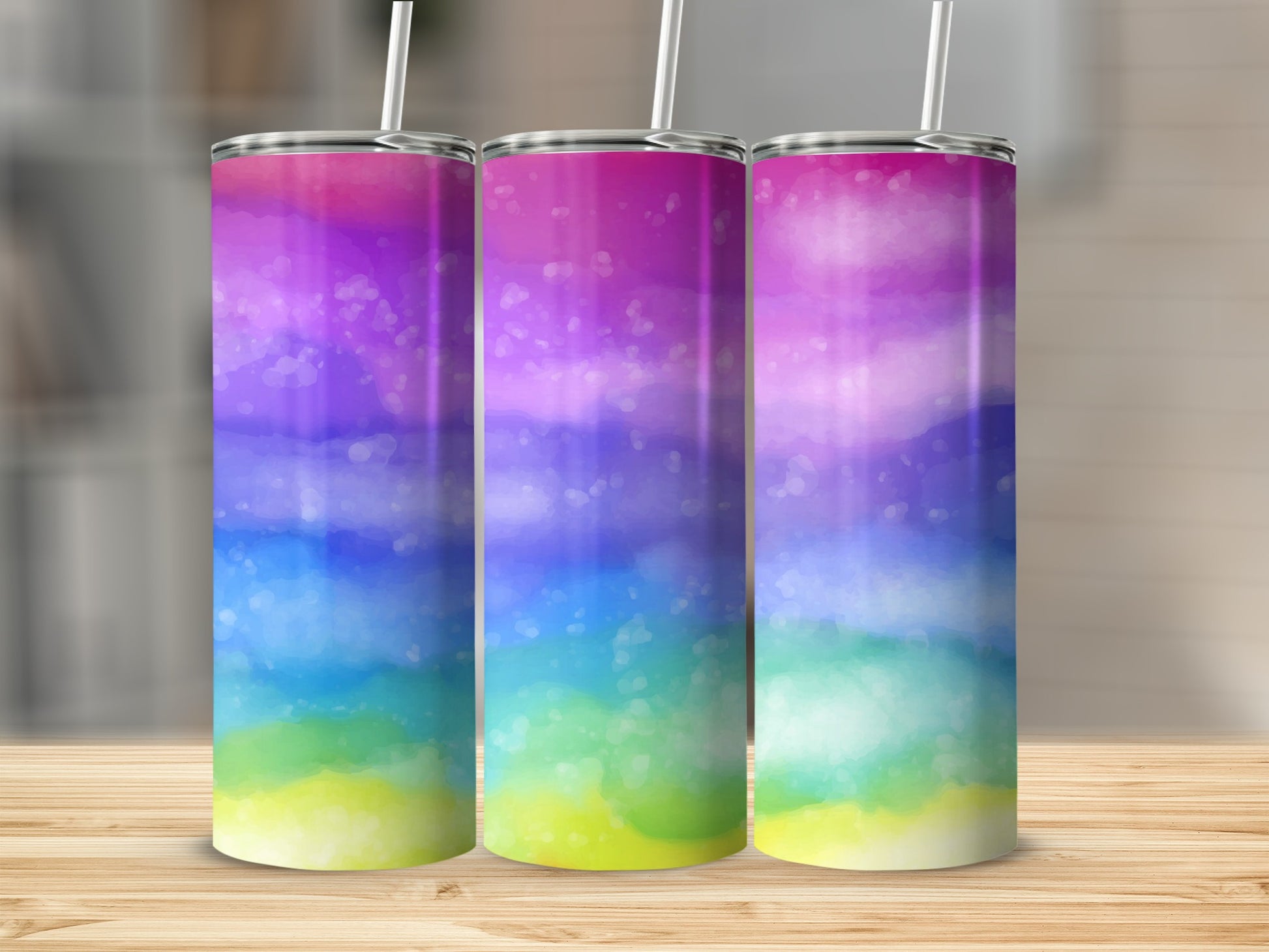 Three tumblers with a watercolor rainbow design and white straws on a wooden surface.