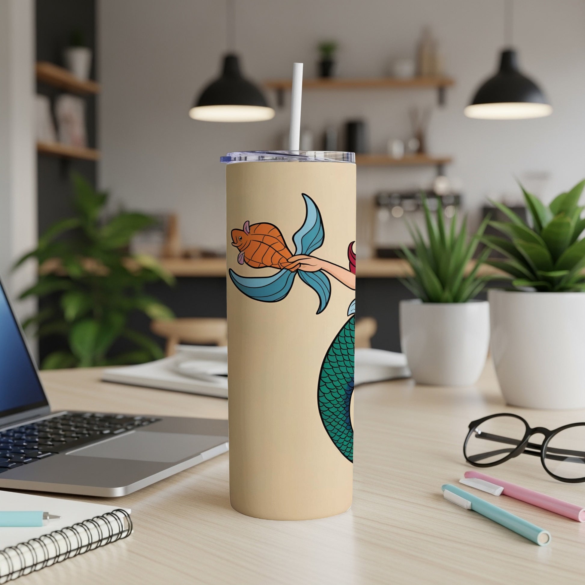 Beige tumbler with cartoon mermaid holding a fish, on a desk.