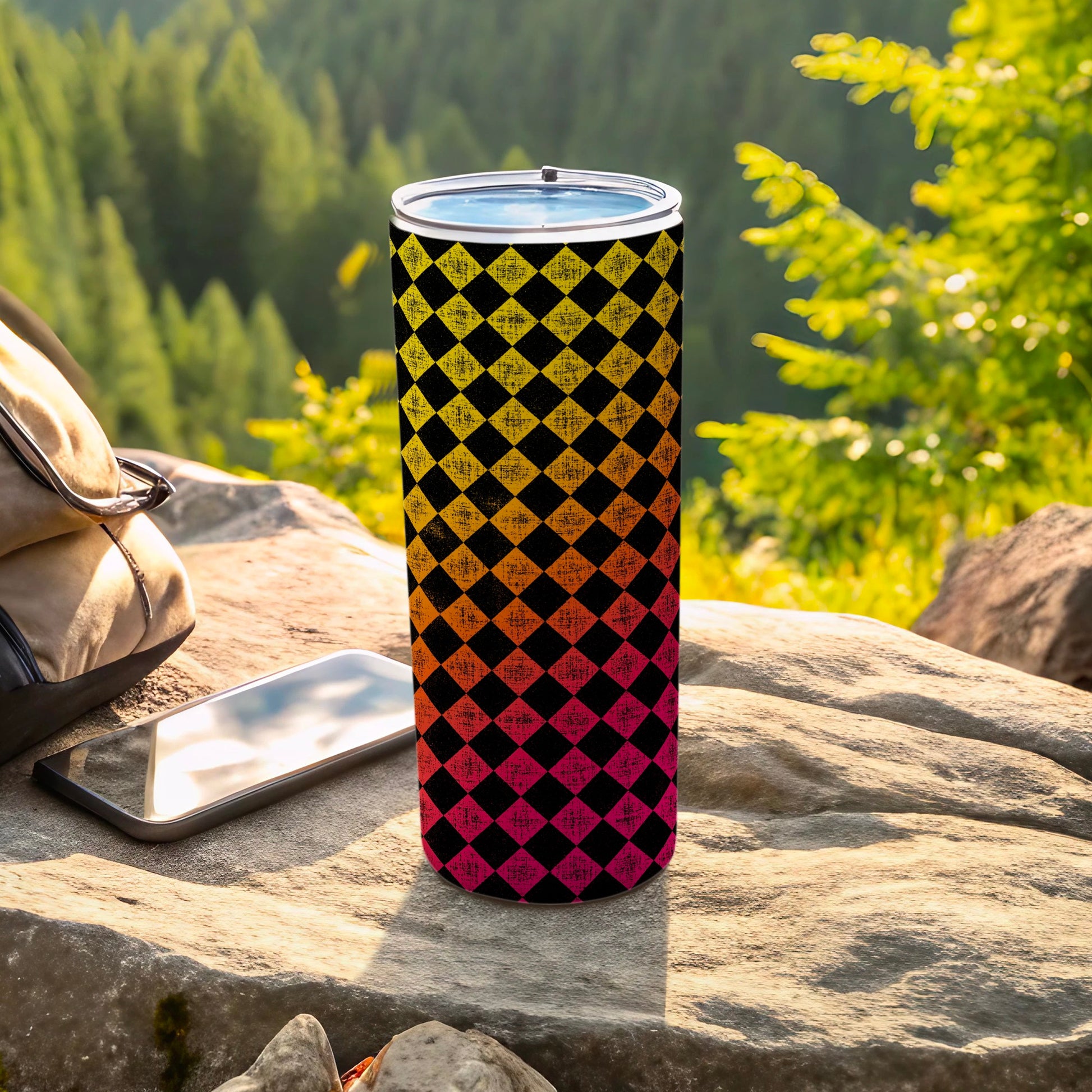 Black, yellow, and orange checkered tumbler on a rock with a backpack and phone.