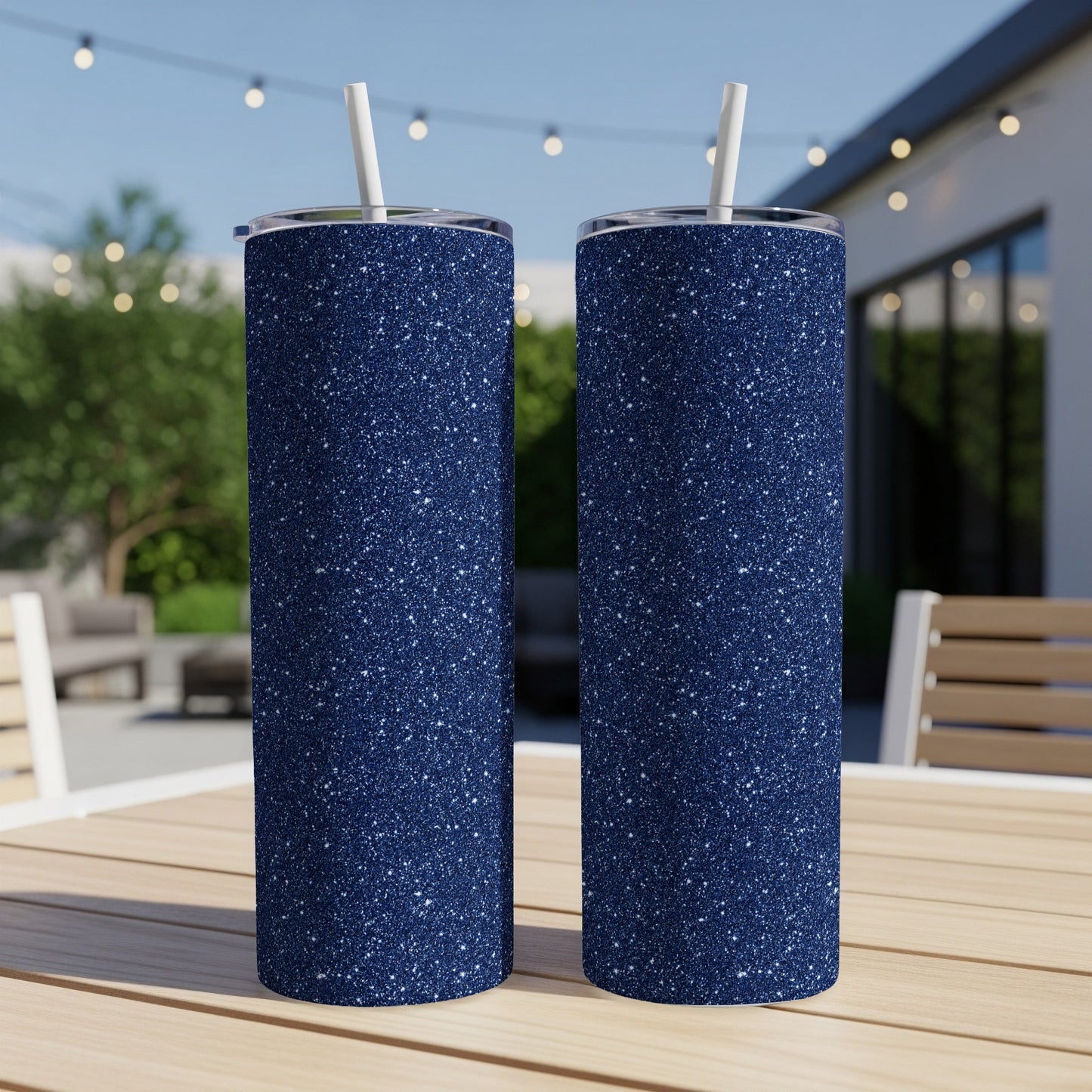 Two navy blue glitter tumblers with white straws sit on a wooden table outdoors.
