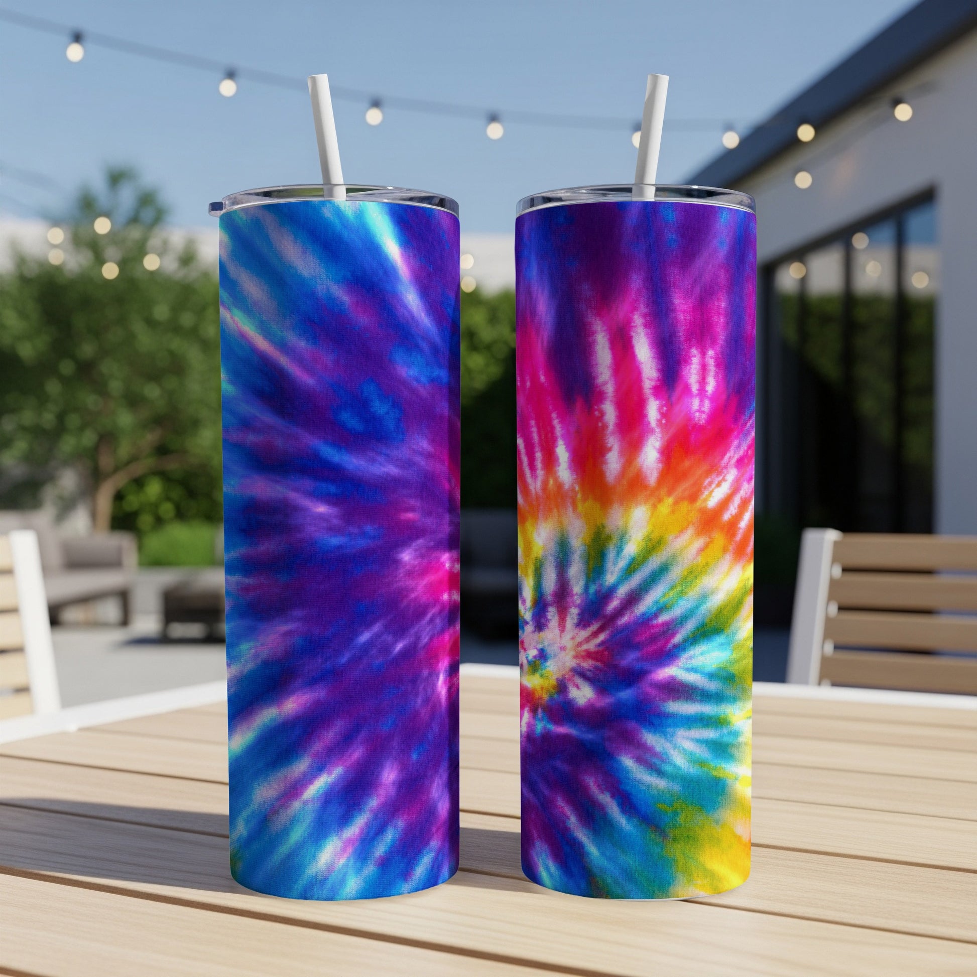 Two tie-dye tumblers with white straws on a wooden table outdoors.