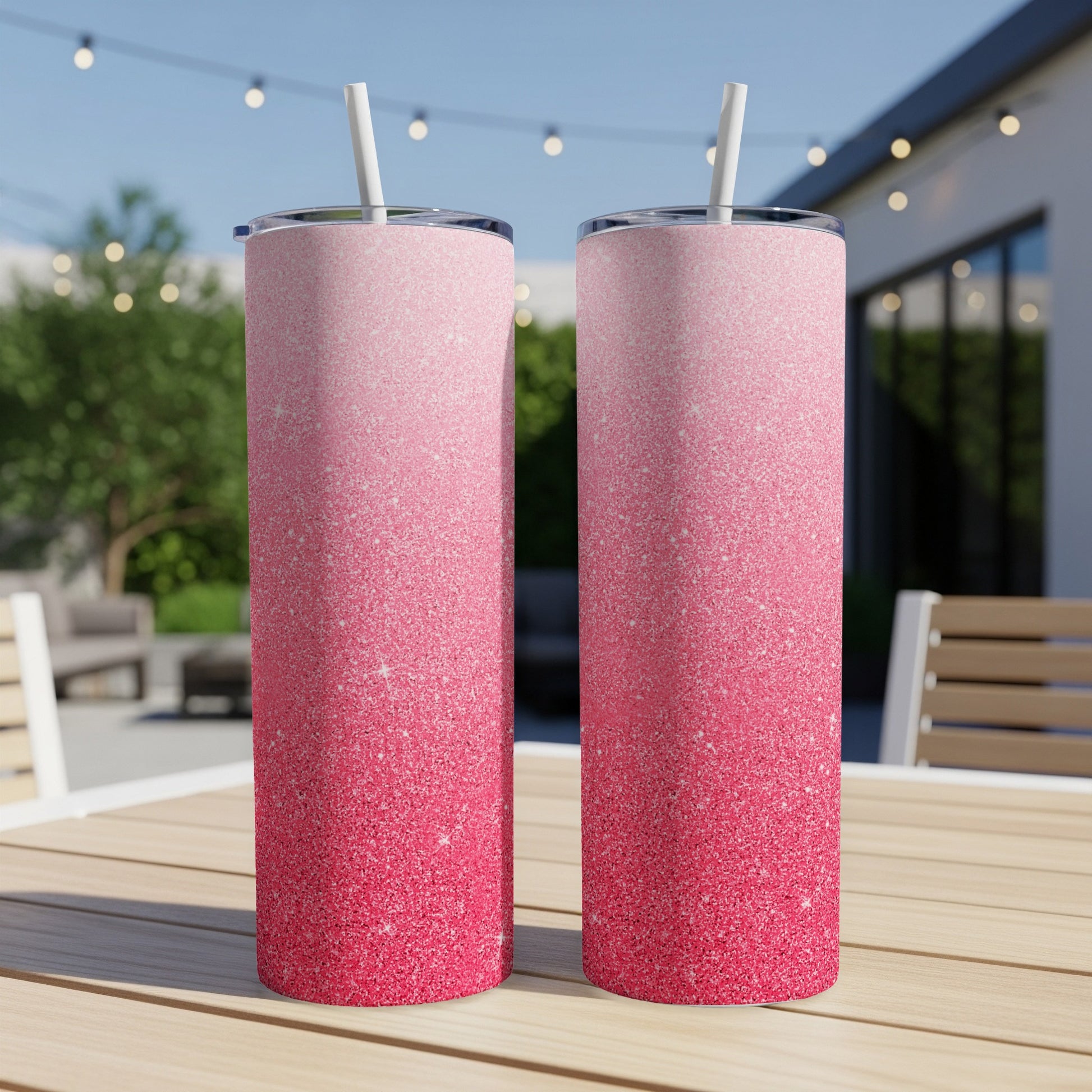 Two pink glitter tumblers with straws on a wooden table outdoors.