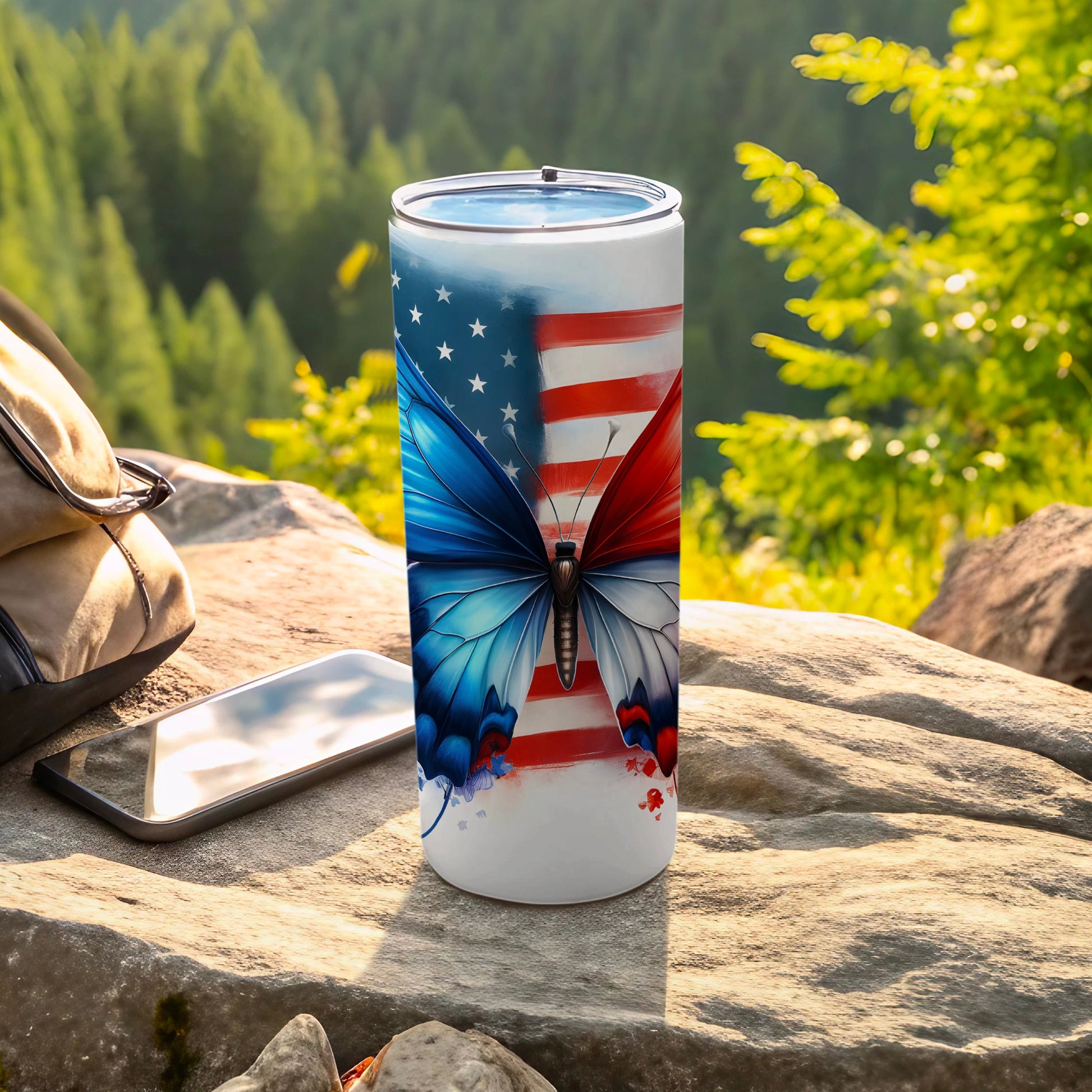Tumbler with American flag butterfly design on rocky surface with phone and backpack.