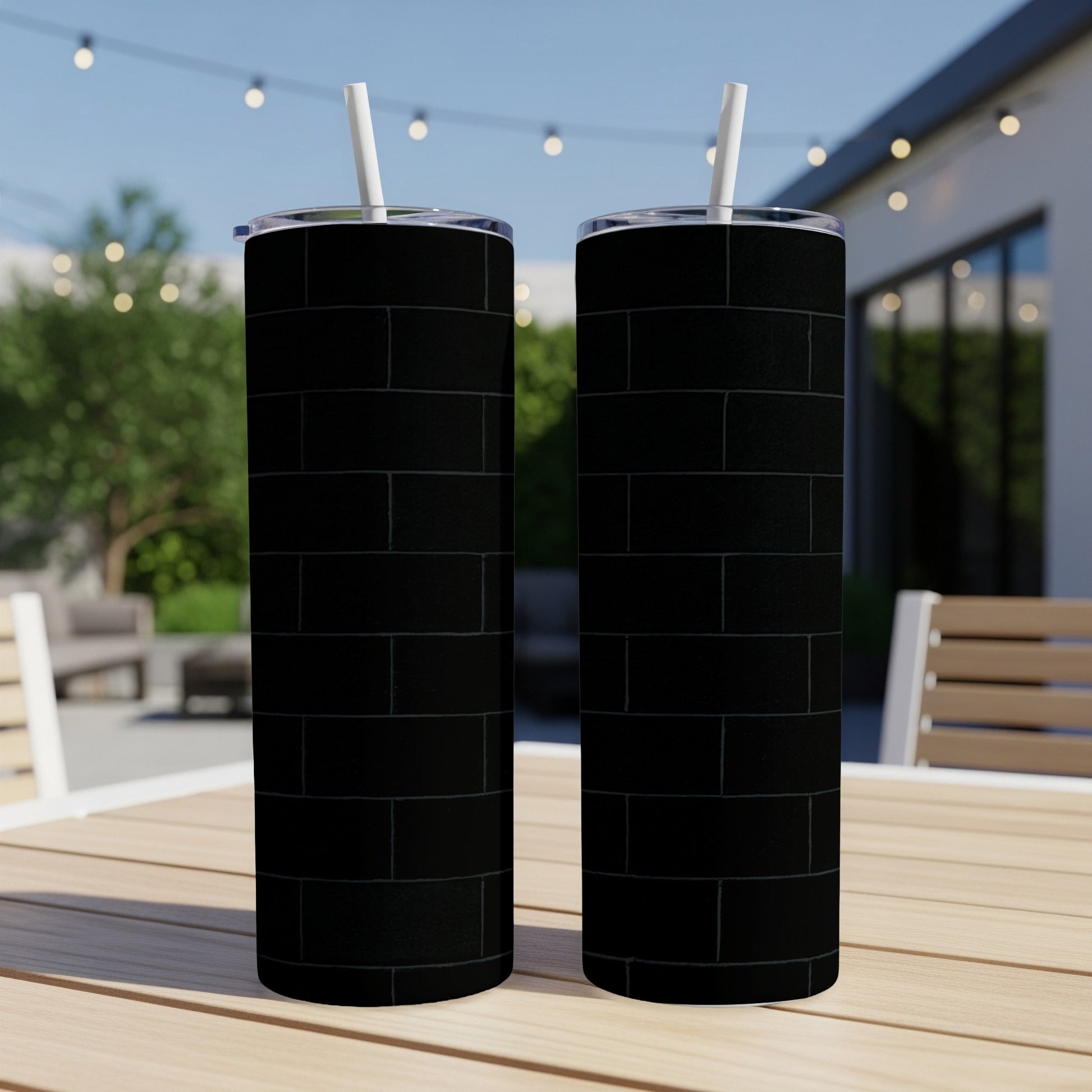 Two black brick pattern tumblers with white straws on a wooden table outdoors.