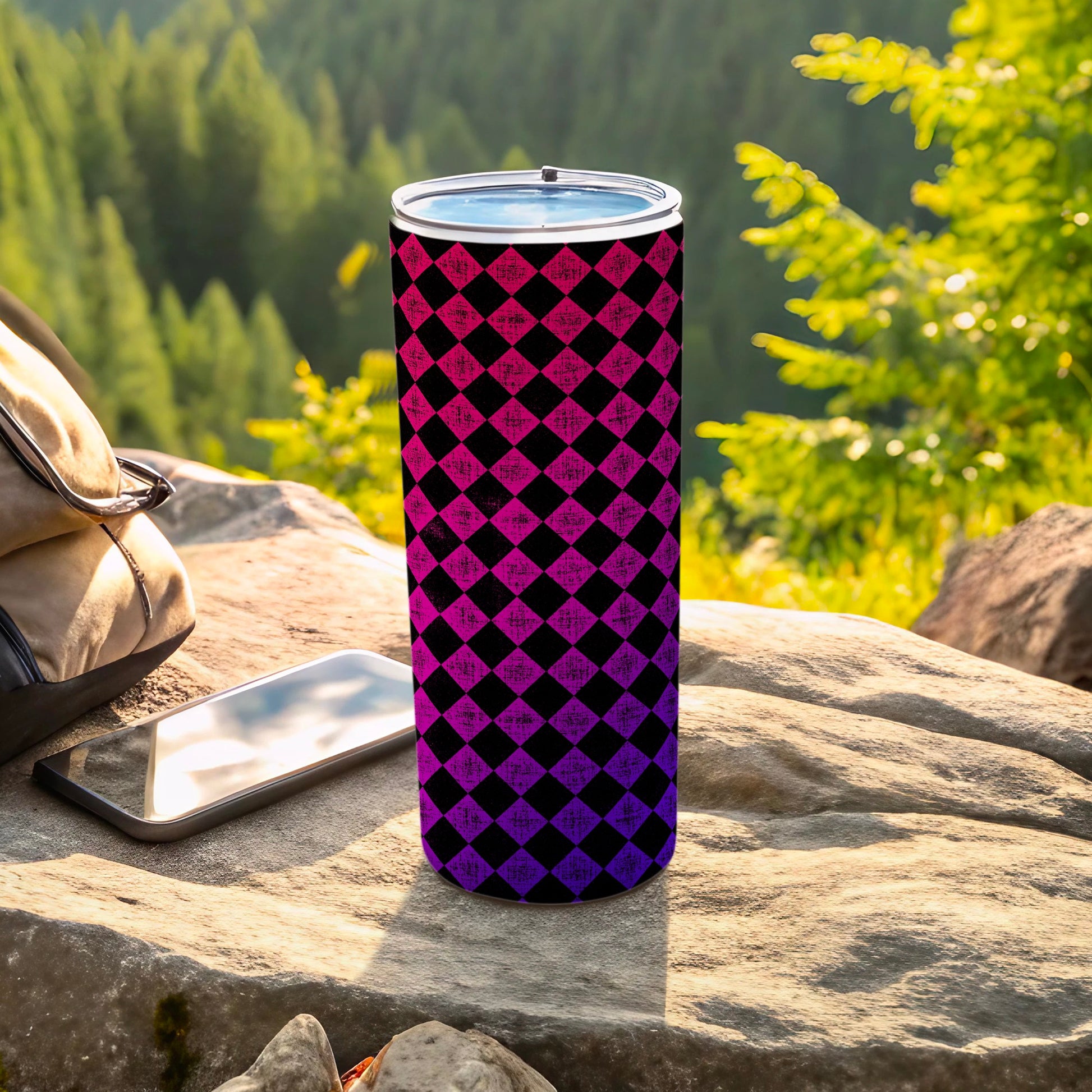 Black and purple checkered tumbler on rocks with backpack and phone.