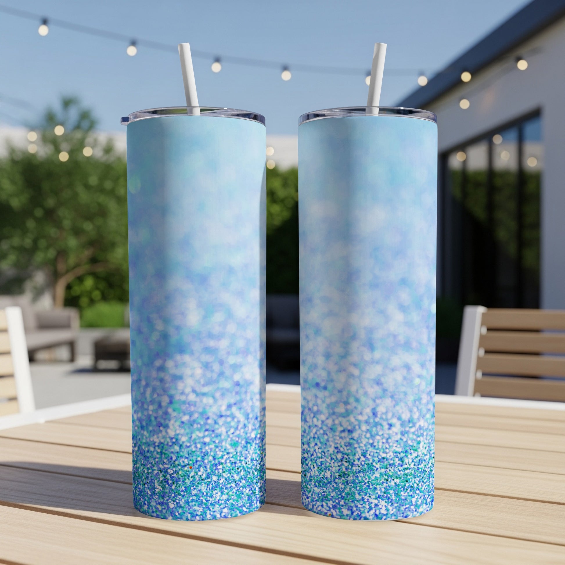 Two tall, blue glitter tumblers with straws on a wooden table outdoors.