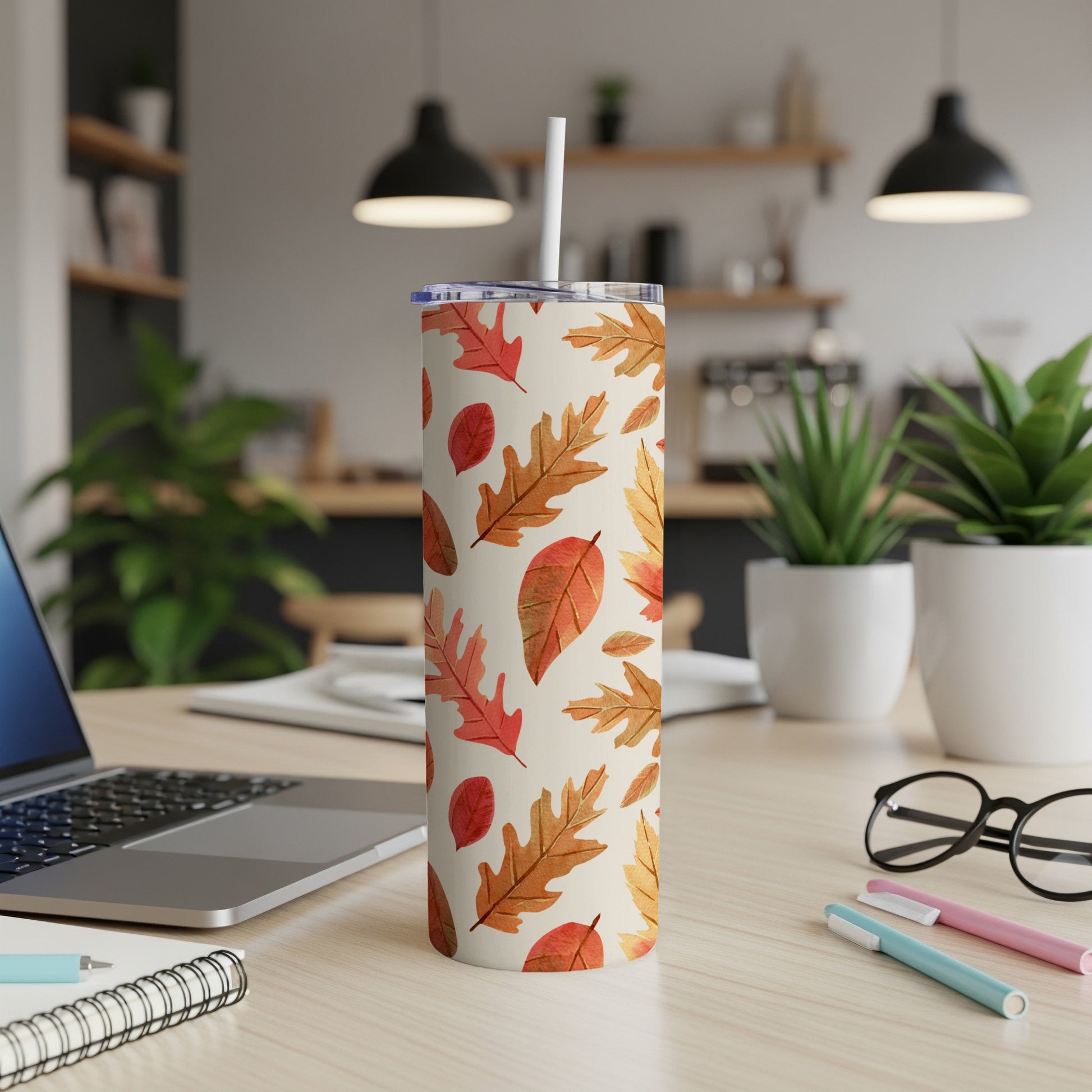 Tan tumbler with autumn leaves pattern and straw, on a desk.