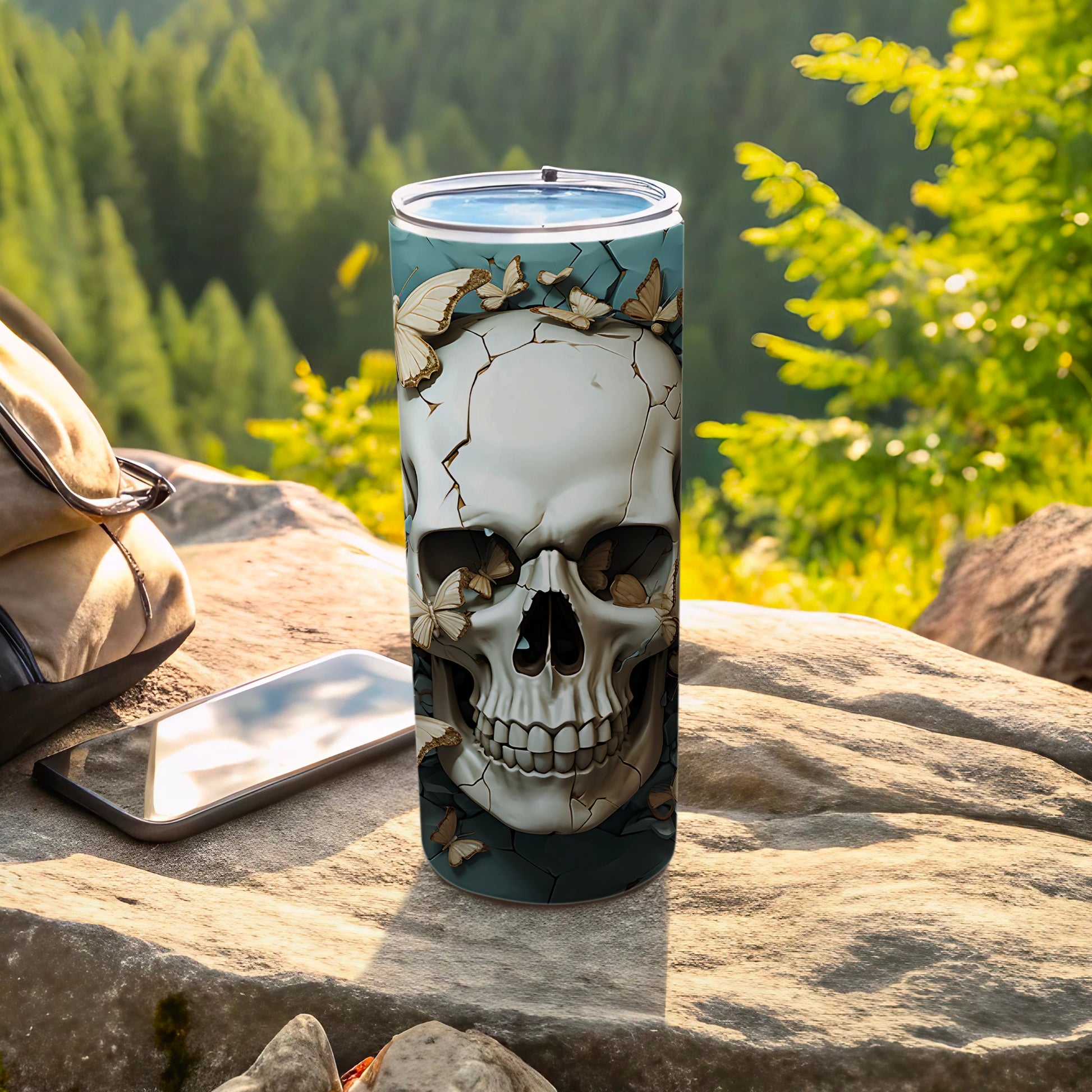 Skull tumbler with butterflies, beside a phone and backpack on rocks.