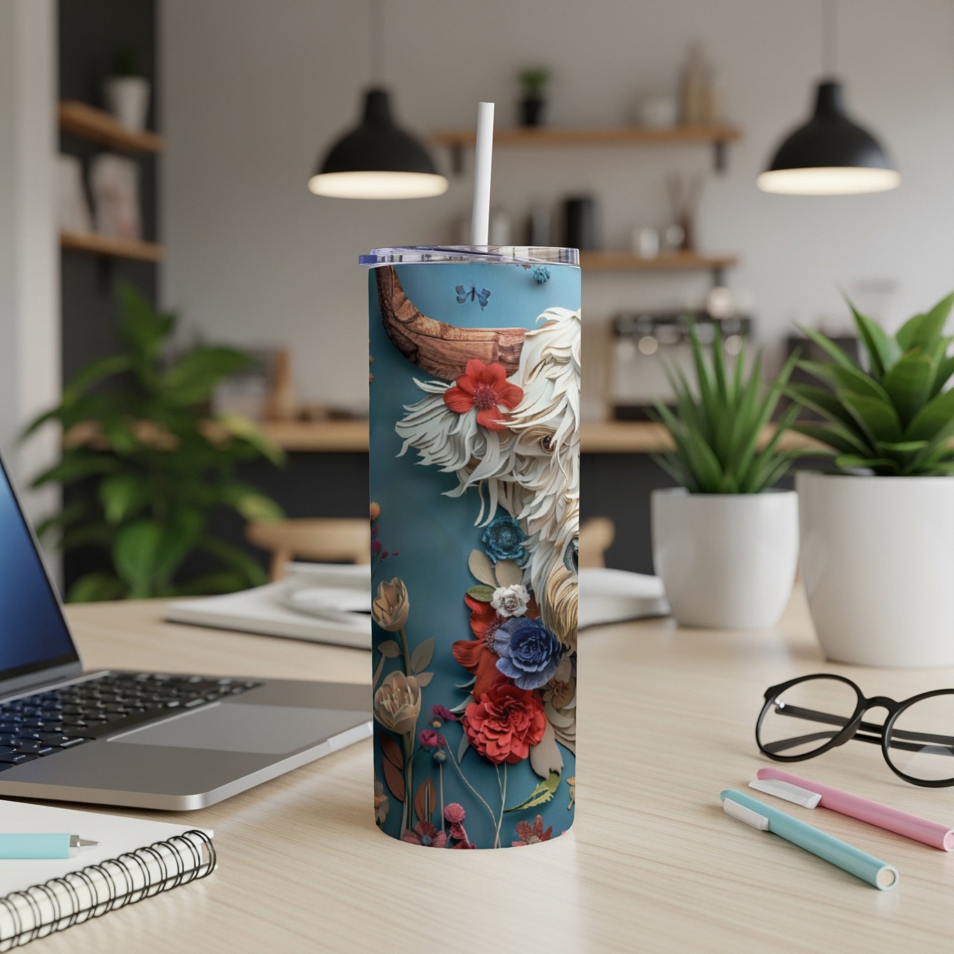 Blue tumbler with floral Highland cow design, straw, and laptop on desk.