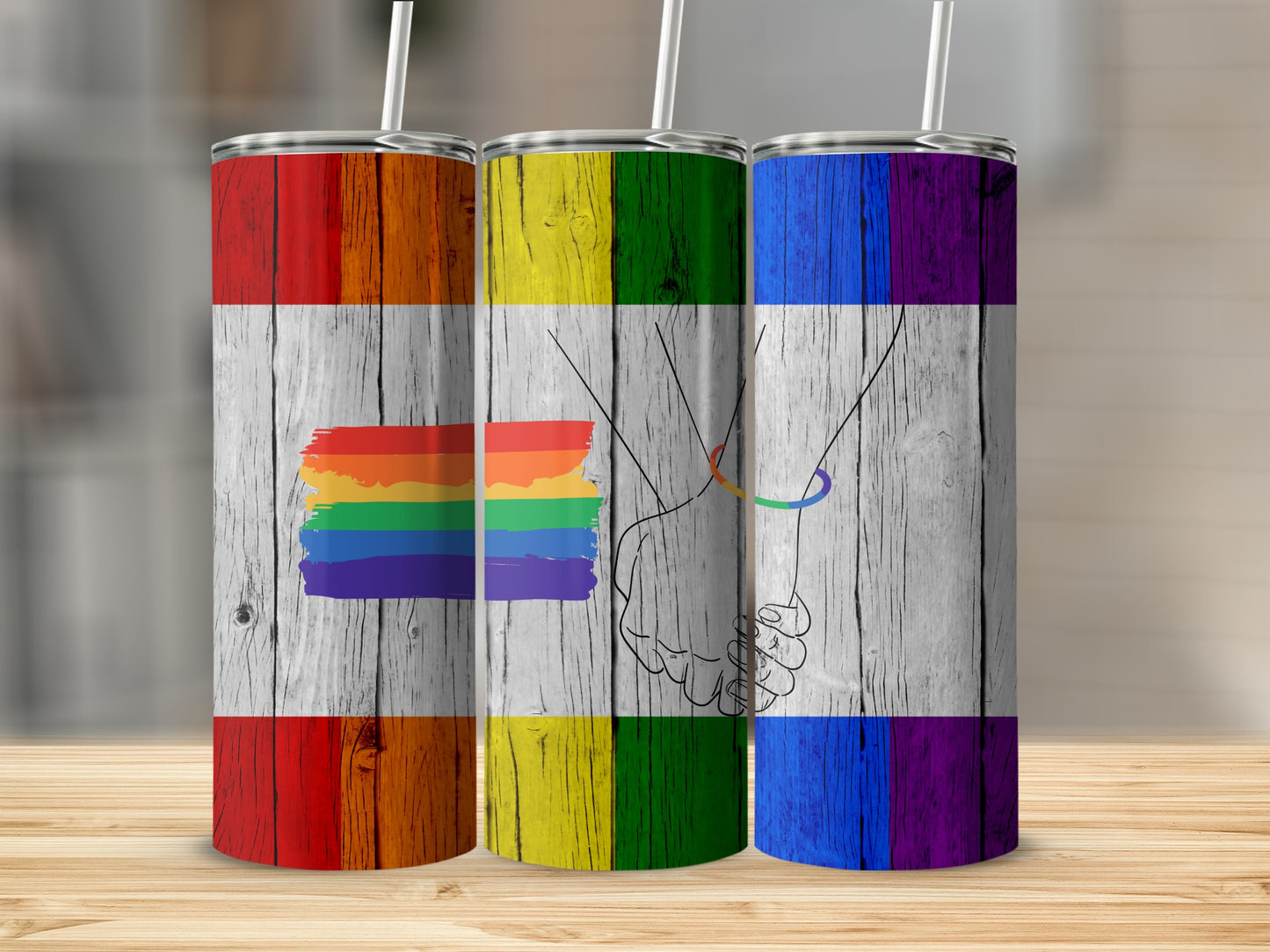 Three tumblers with rainbow flag design on wood grain background.