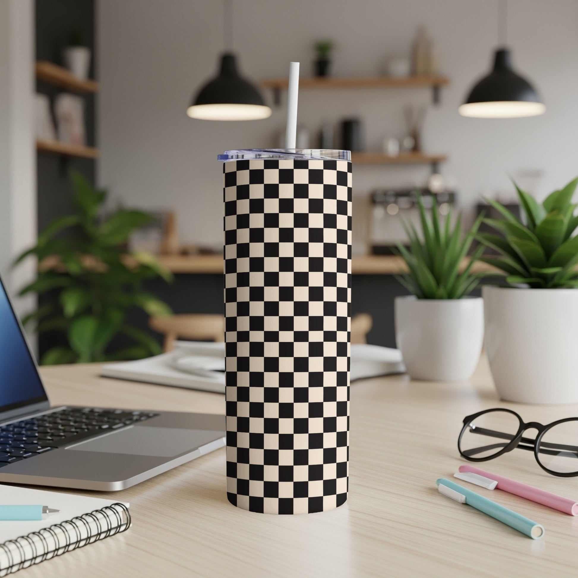 Checkerboard tumbler with a straw on a wooden desk with a laptop.