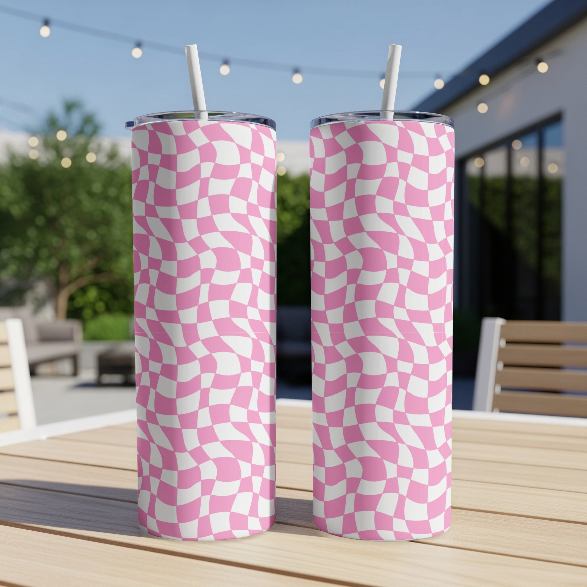 Two pink and white checkered tumblers with straws sit on a wooden table outdoors.