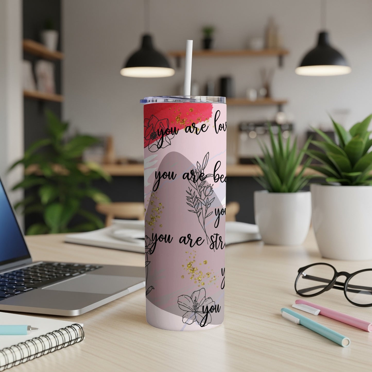 Pink tumbler with text "you are loved" and floral accents on a desk.
