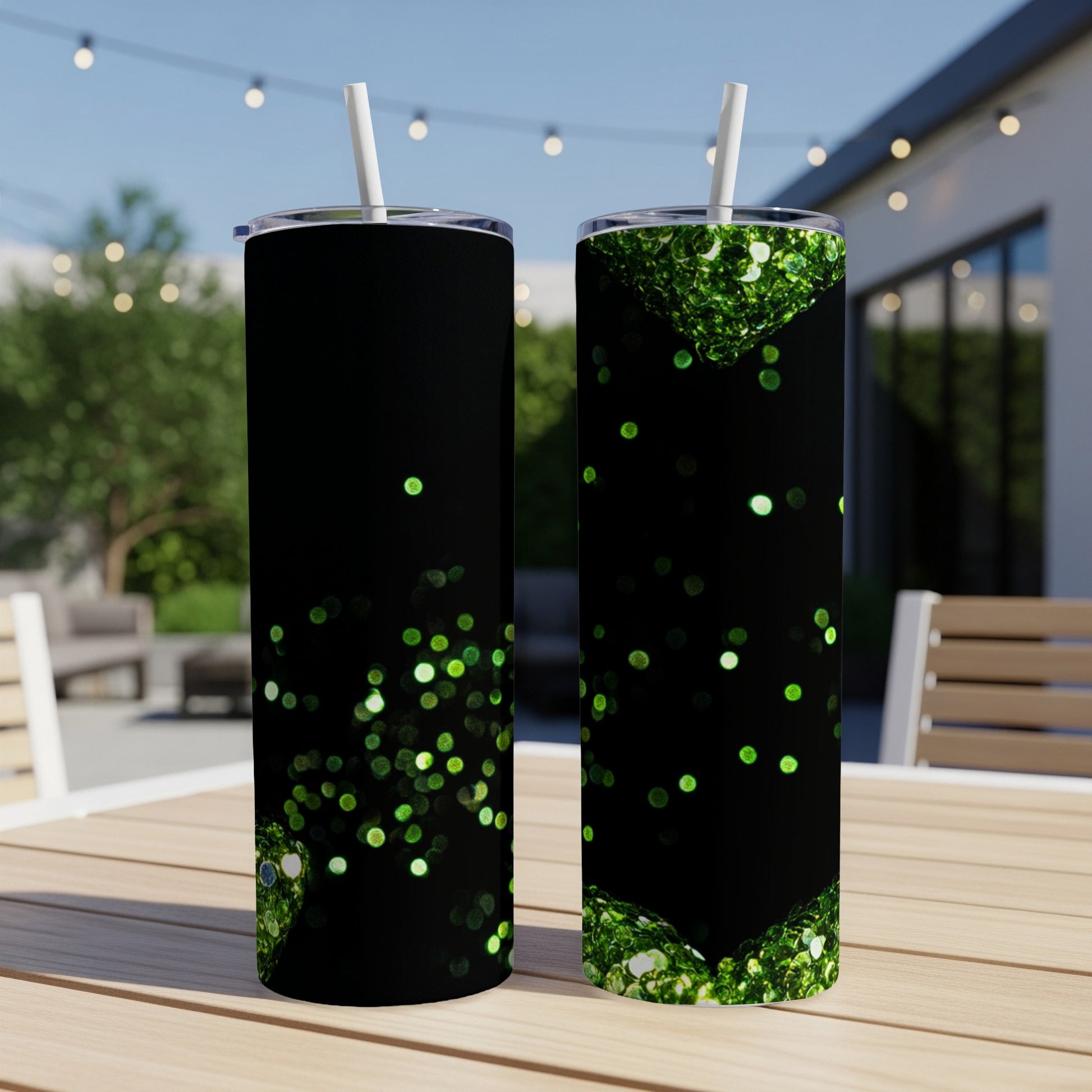 Two black tumblers with green glitter designs and straws on a patio table.