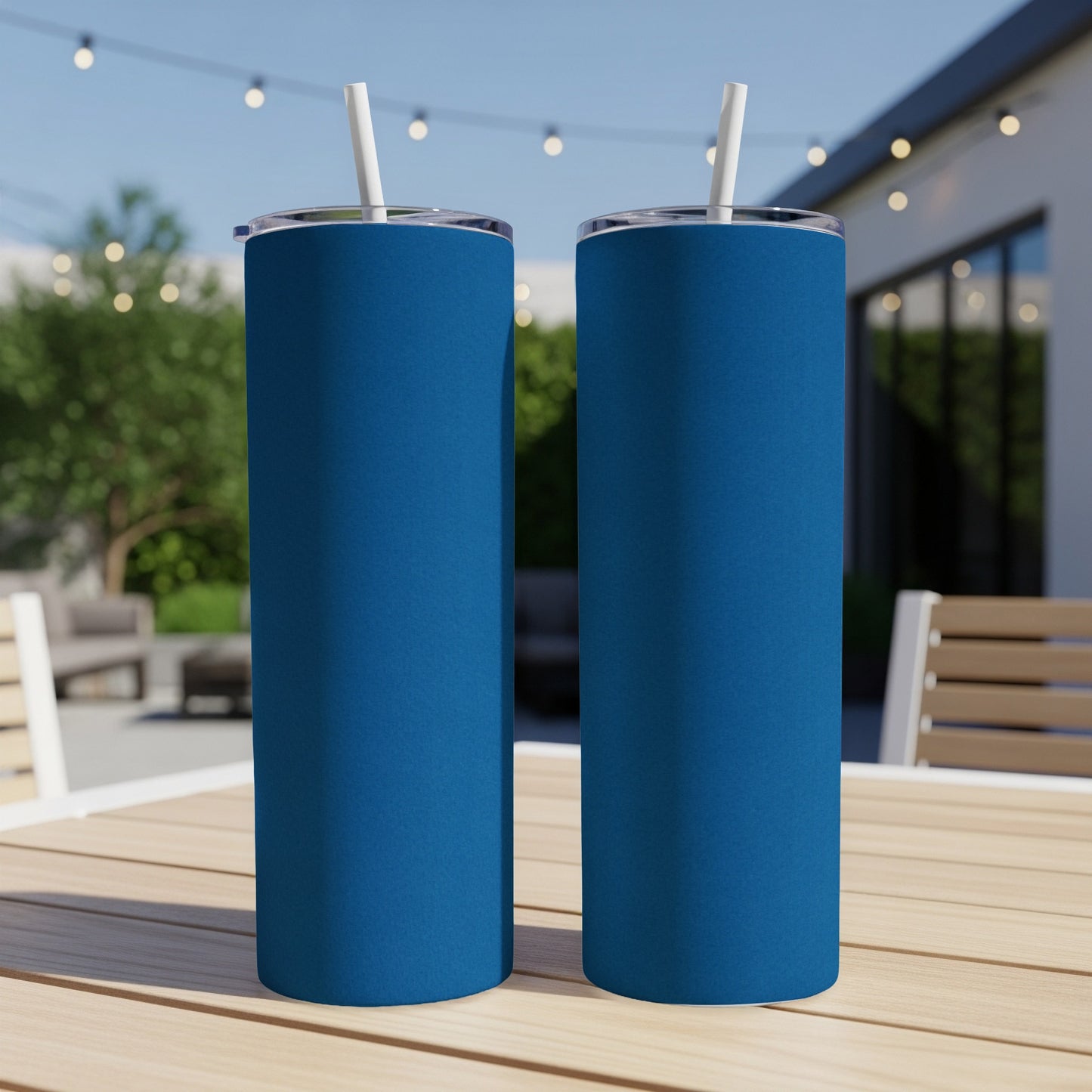 Two matte blue tumblers with white straws sit on a wooden table outdoors.