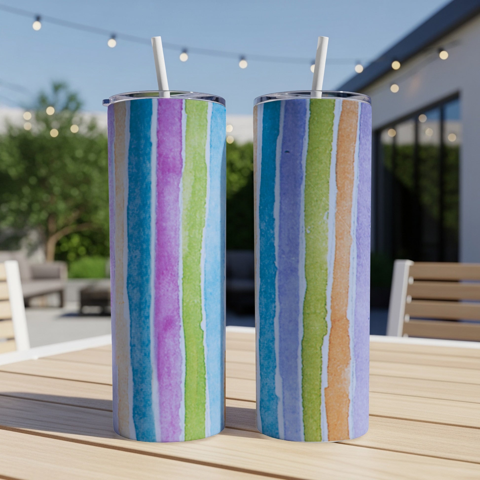 Two colorful striped tumblers with straws on a wooden table outdoors.