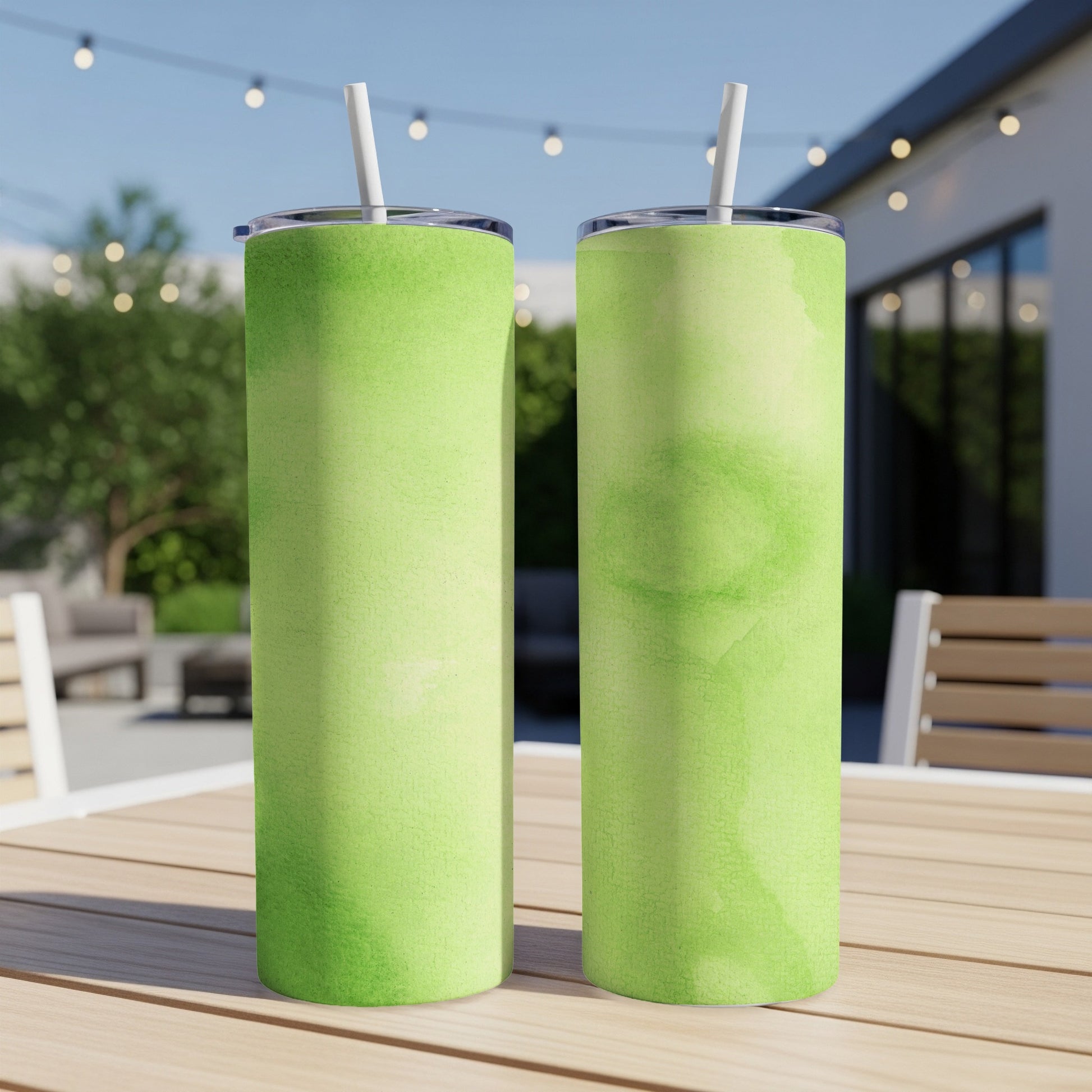 Two green watercolor patterned tumblers with straws on a wooden table outdoors.