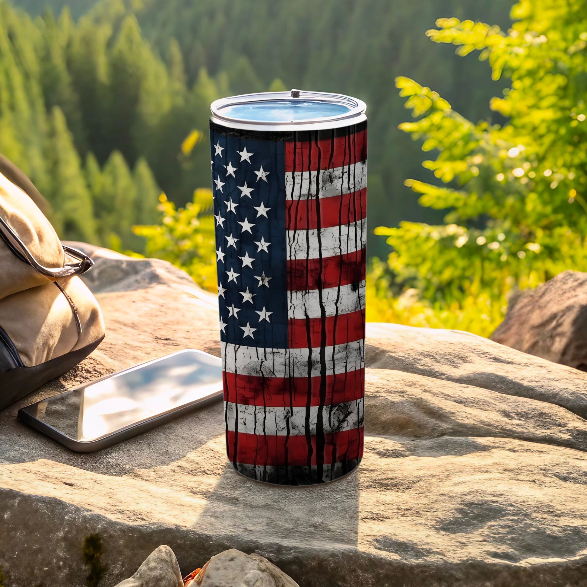 Distressed American flag tumbler on rock with backpack and phone.