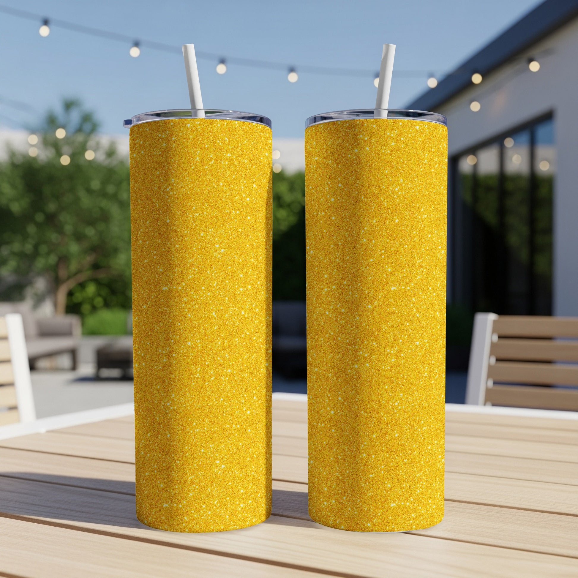 Two glittery gold tumblers with straws sit on a wooden table outdoors.