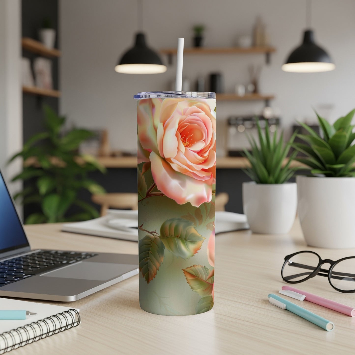 Floral rose tumbler with a straw on a desk with a laptop and pens.