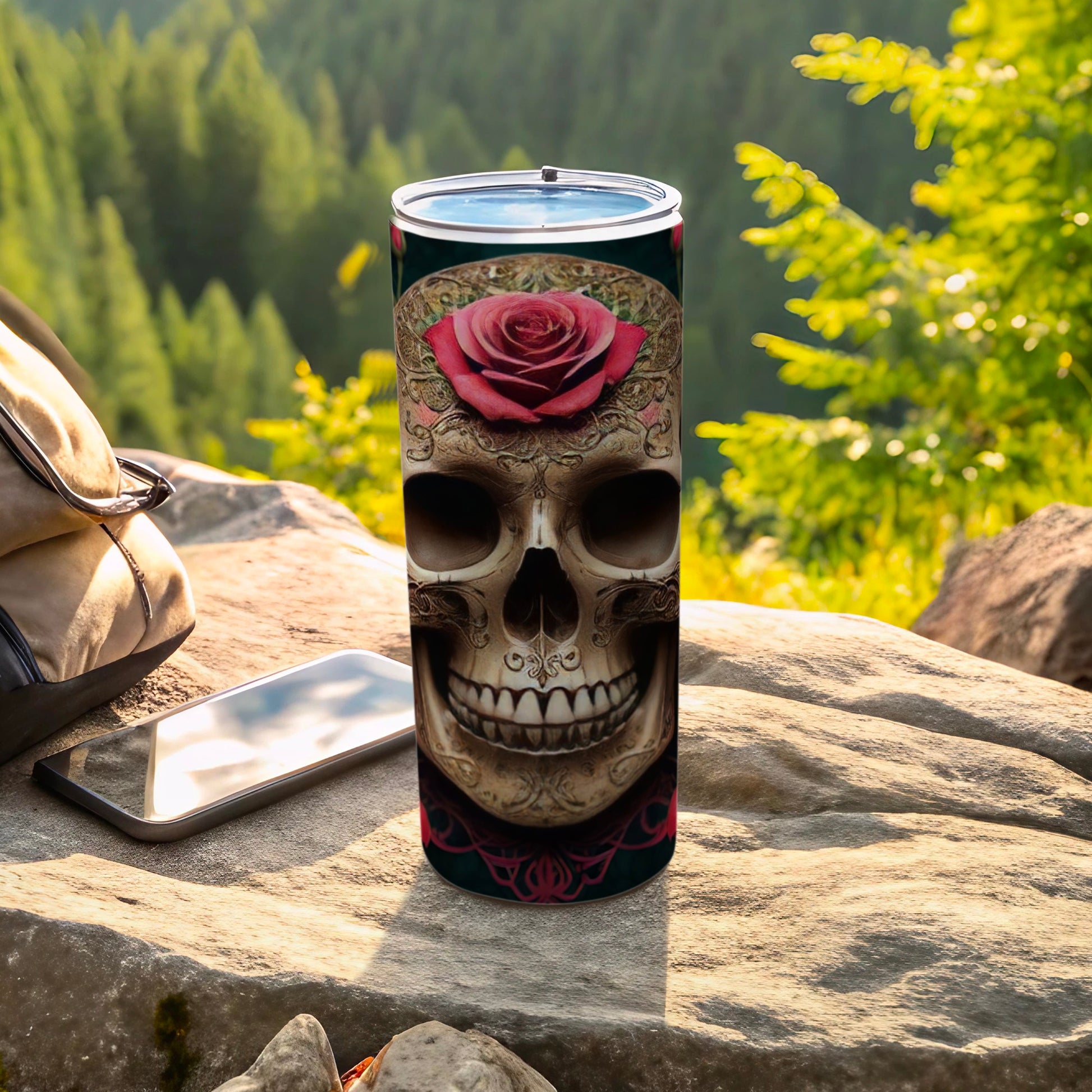 Skull with rose design on a tumbler, beside a phone and bag.