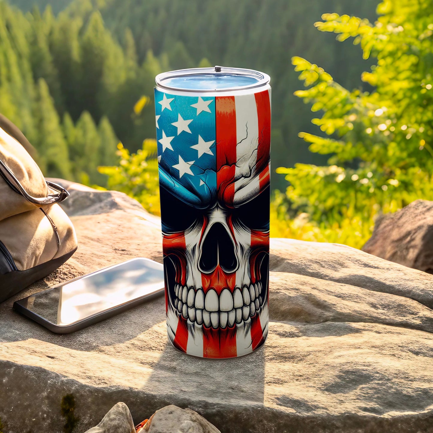 Tumbler with American flag skull design, resting on rocks with phone and backpack.